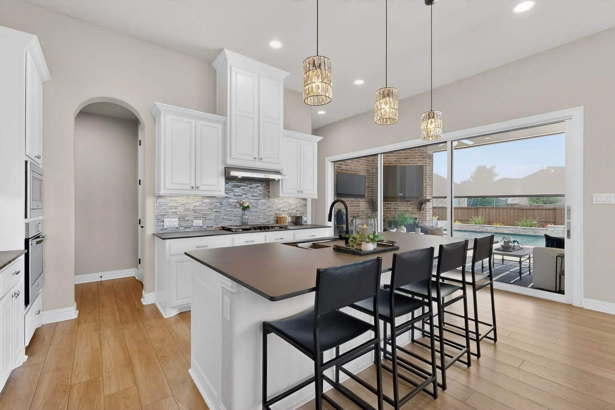 Modern kitchen and dining area featuring white cabinetry, a quartz countertop island with seating for three, stainless steel appliances, subway tile backsplash, pendant lighting, wood-finish flooring, and expansive sliding glass doors opening to an outdoor entertaining space with countryside backdrop