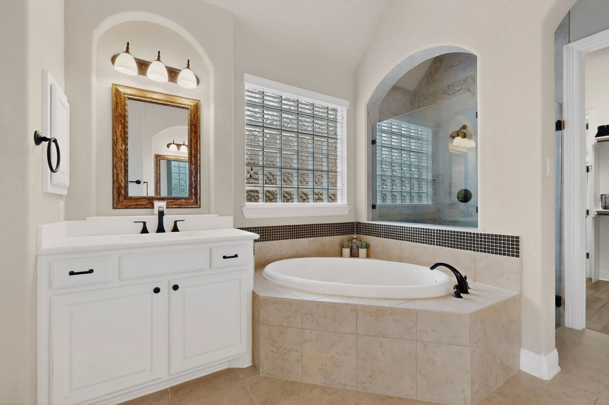 White vanity with black hardware, arched wall niche, built-in bathtub with tiled surround, glass block window, and frameless glass shower enclosure