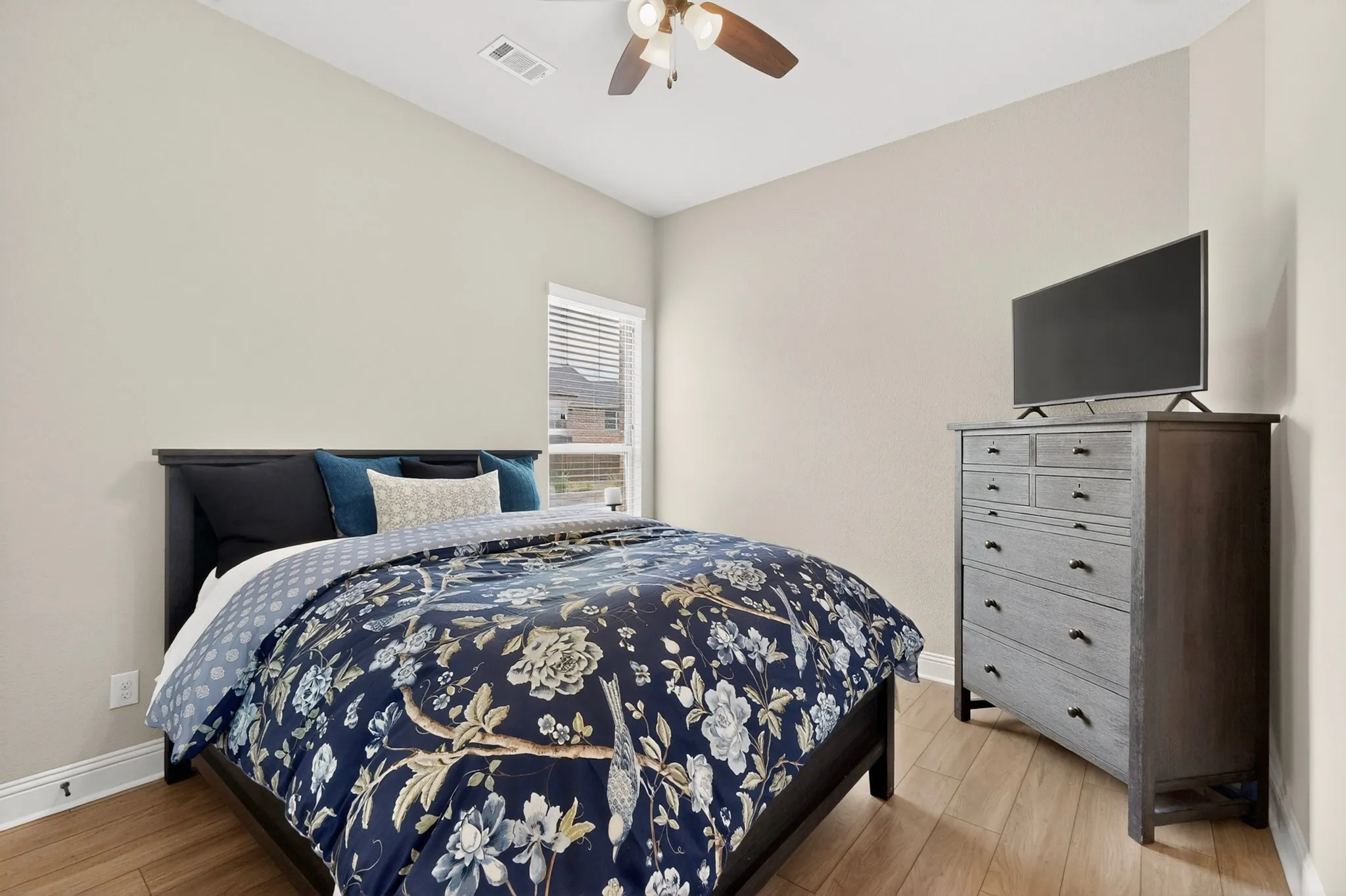 Spacious bedroom featuring warm wood-finish flooring, a ceiling fan with integrated lighting, and ample natural light from a louvered window