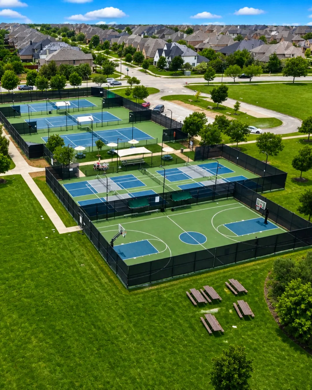 Recreational area featuring multiple fenced tennis courts, additional fenced sports courts, a dedicated basketball court, and picnic tables on a grassy expanse