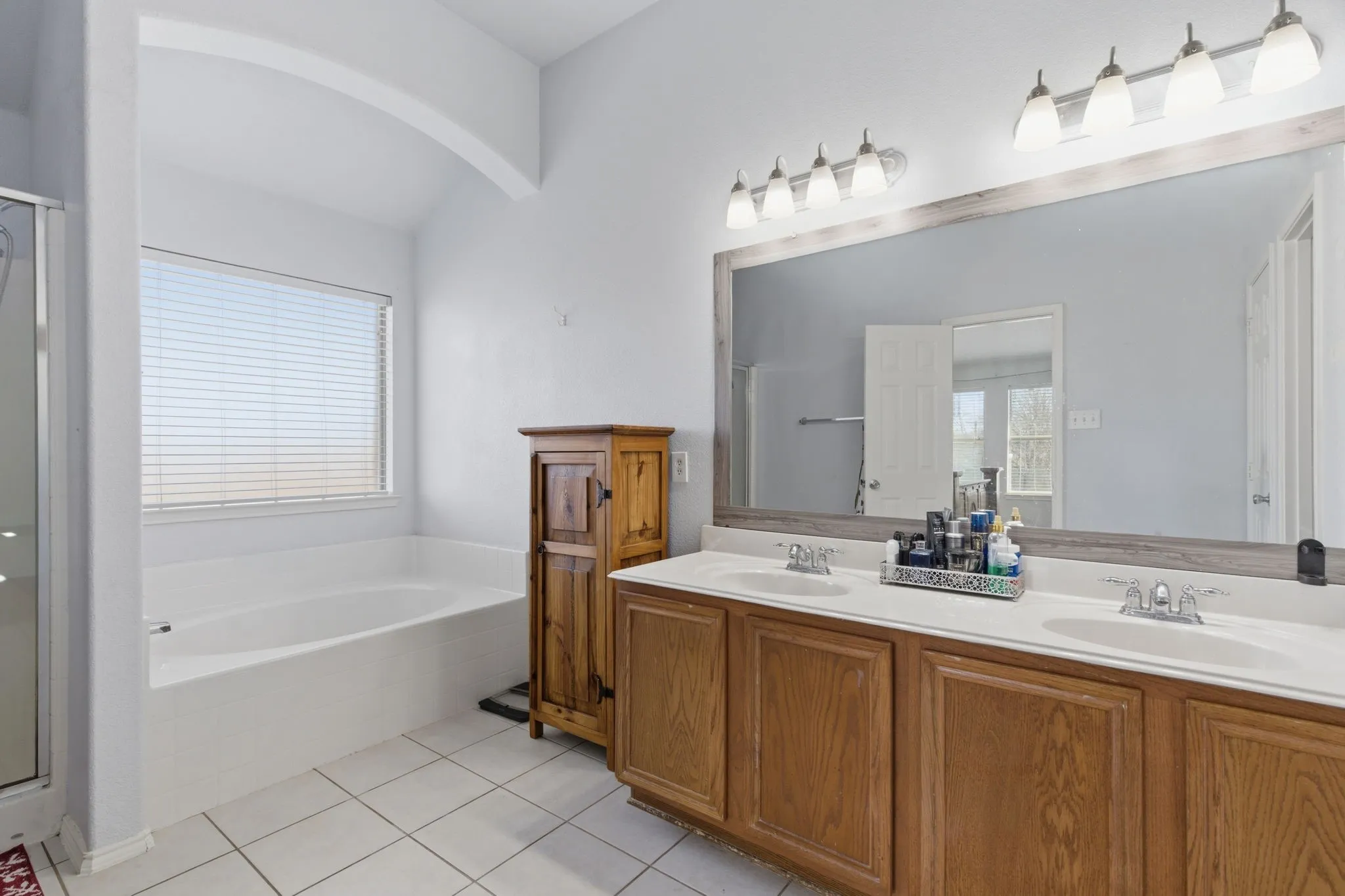 Bathroom featuring a white soaking tub under a large window with blinds, dual basin vanity with wood cabinetry, and a framed mirror