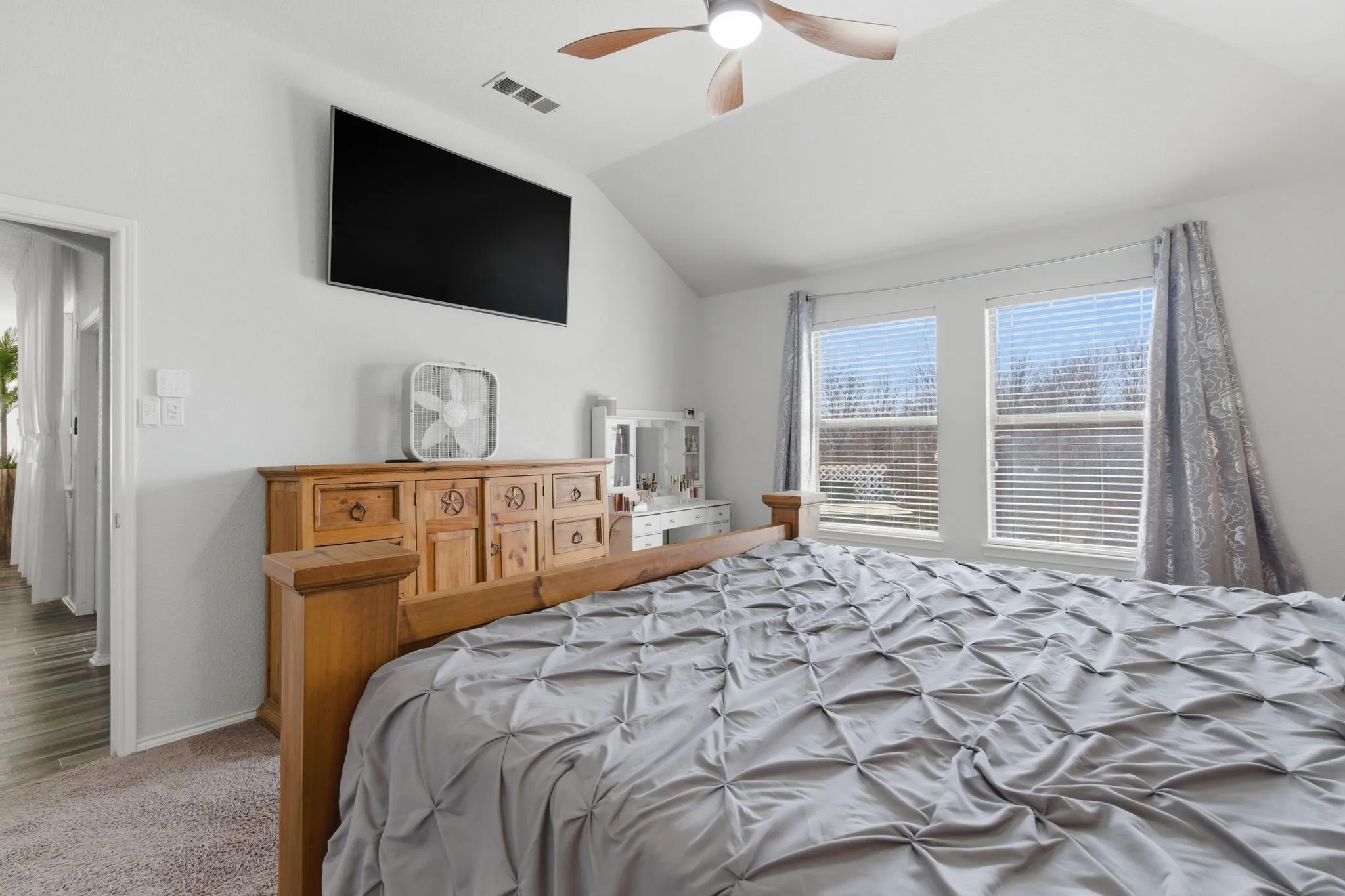 Vaulted ceiling bedroom featuring plush carpeting, dual windows with blinds, a wood-finish ceiling fan, and a wall-mounted flat-screen display