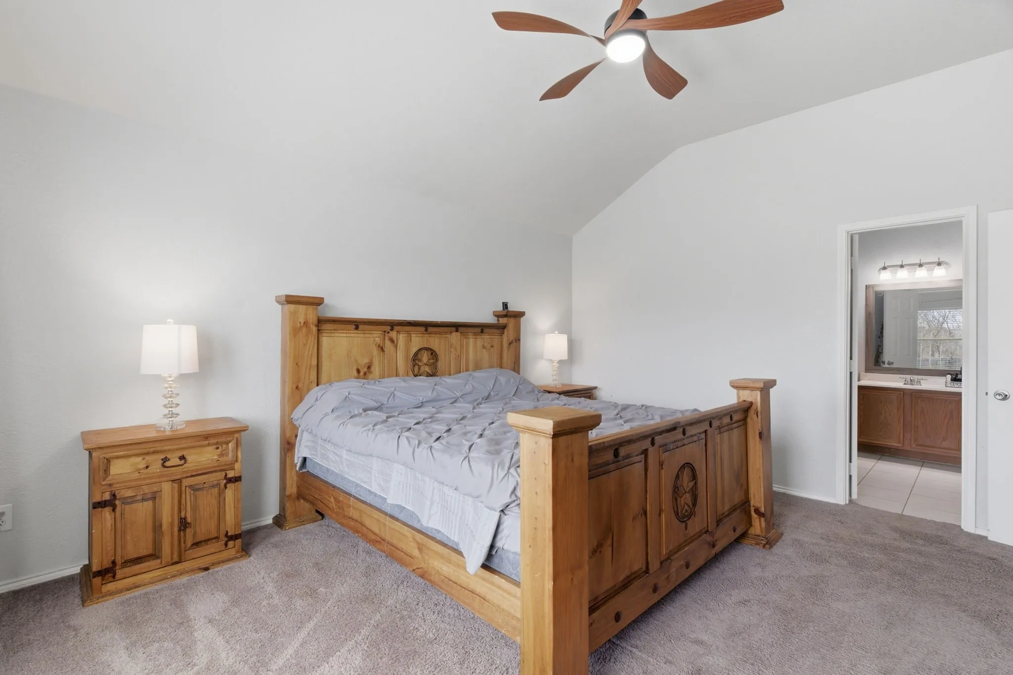 Carpeted room featuring a ceiling fan with light fixture, vaulted ceiling, and an open doorway revealing a bathroom with a vanity and mirror