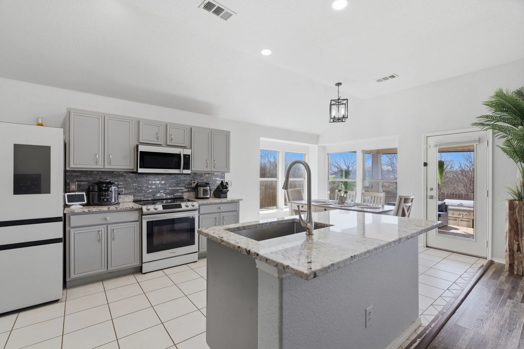 Open-concept kitchen featuring an island with an undermount sink, granite countertops, gray cabinetry, stainless steel appliances, and a tile backsplash