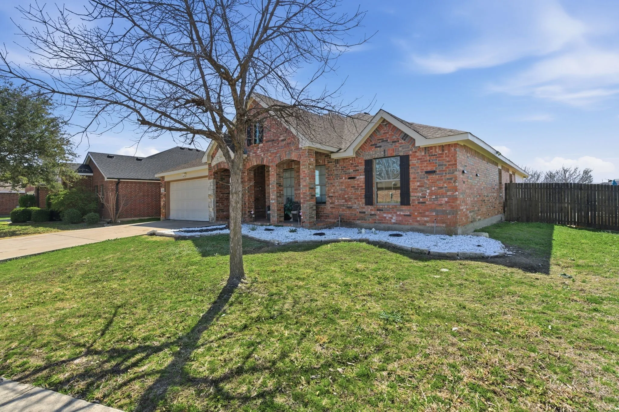 Brick exterior home featuring an attached garage, arched entry, front yard with mature tree, and a white rock landscape bed
