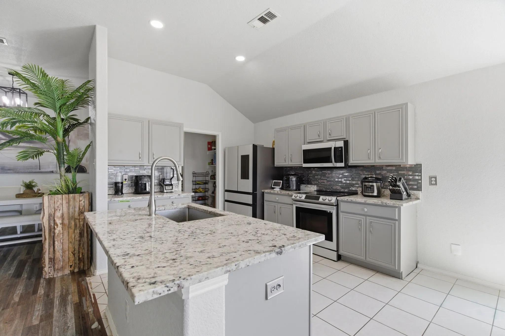 Kitchen featuring a center island with a stainless steel sink, granite-finish countertops, light gray cabinetry, and a dark tile backsplash