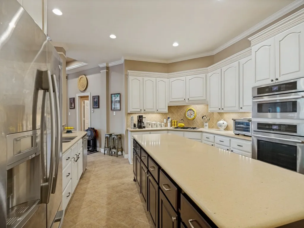 Kitchen featuring an extended island with a light-toned countertop, white upper cabinetry, stainless steel double wall ovens, a coordinating stainless steel refrigerator, and recessed lighting