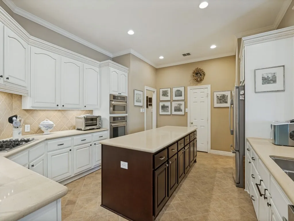 Spacious kitchen featuring an expansive center island, white cabinetry, light-toned countertops, diagonal tile backsplash, and integrated stainless steel double ovens