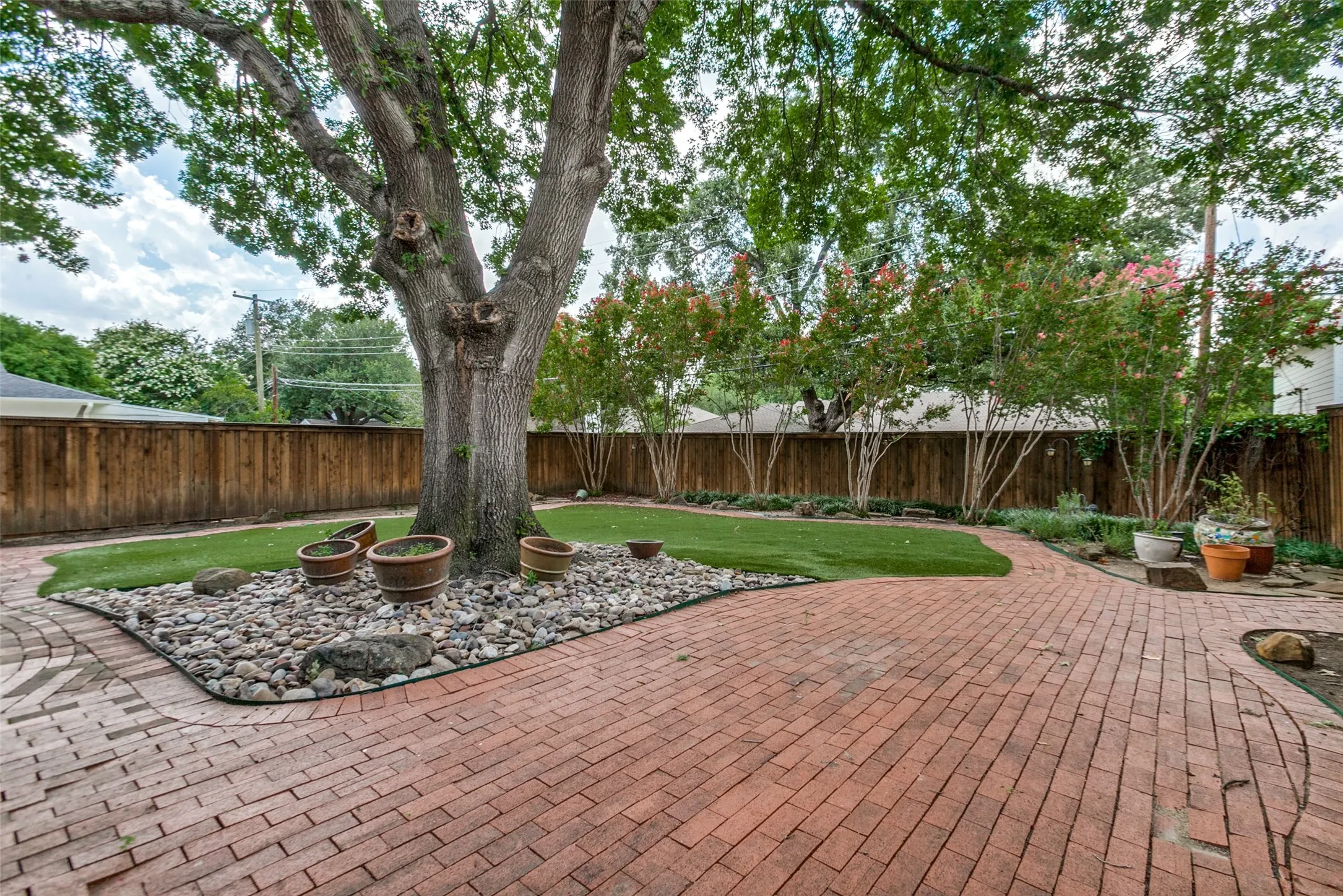 Private backyard featuring extensive red brick paver patio, mature shade tree, and artificial turf lawn