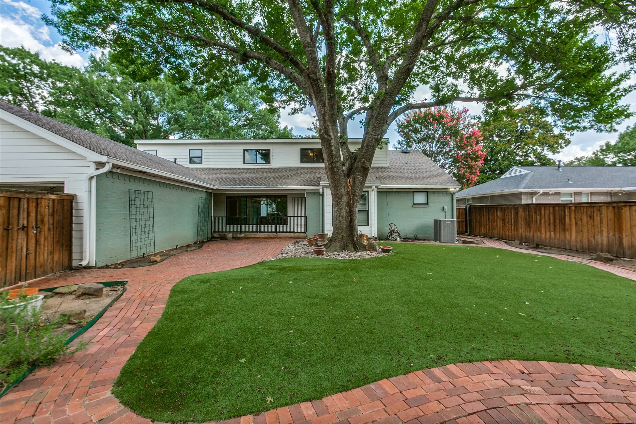 Expansive backyard featuring a manicured lawn, mature shade tree, brick paver patio, and full privacy fencing
