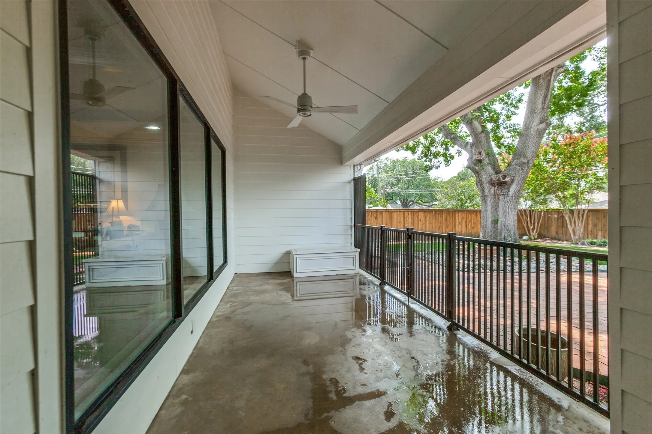Covered outdoor patio features a ceiling fan and a black metal railing overlooking a fenced yard with mature trees