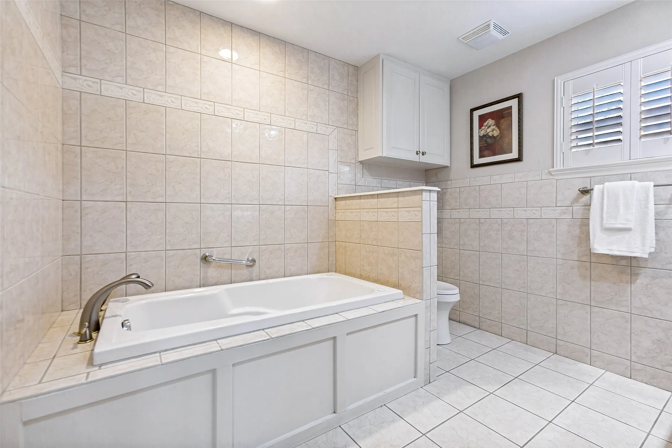 Tiled bathroom featuring a built-in soaking tub with brushed nickel fixtures, an adjacent toilet, and a window with plantation shutters