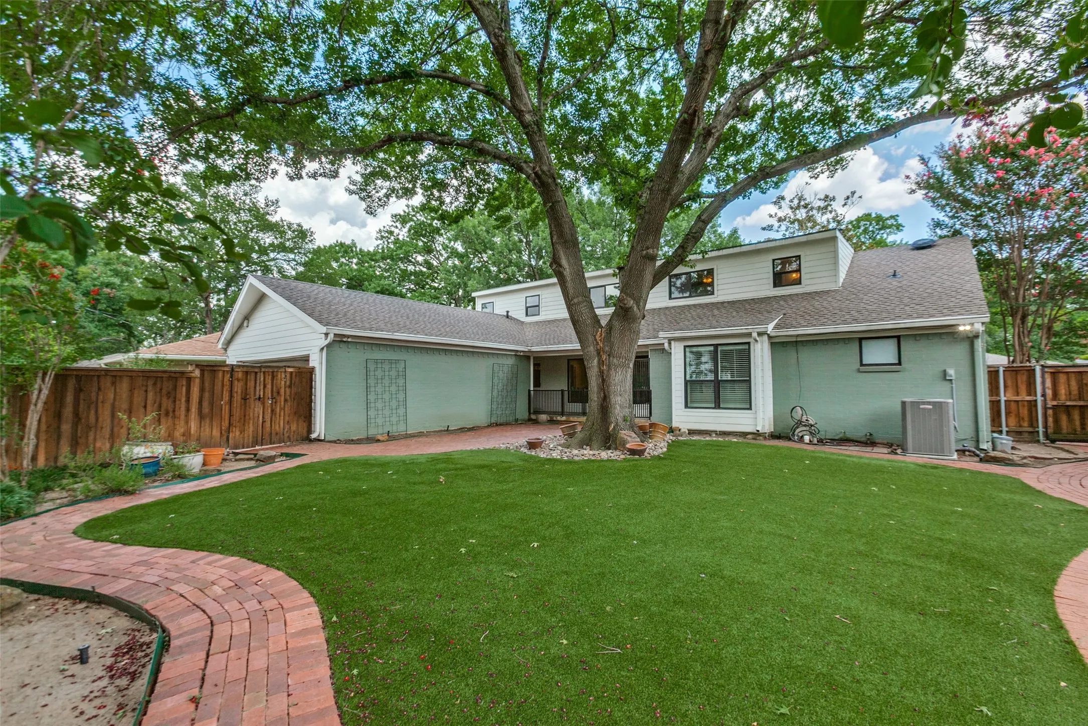 Expansive backyard featuring a large central tree, manicured artificial turf lawn, and brick paver pathways