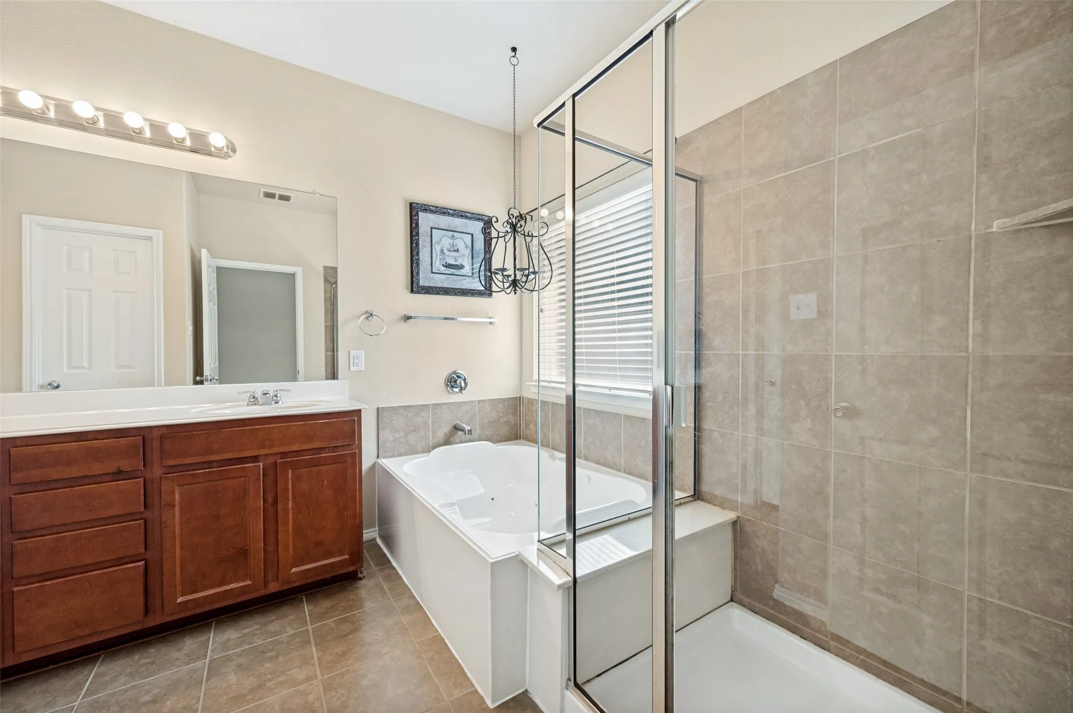 Bathroom featuring a dual-sink vanity with wood-finish cabinetry and white countertop, an enclosed shower with full-height tile surround, a corner soaking tub with tile surround, a window with horizontal blinds, and tile flooring