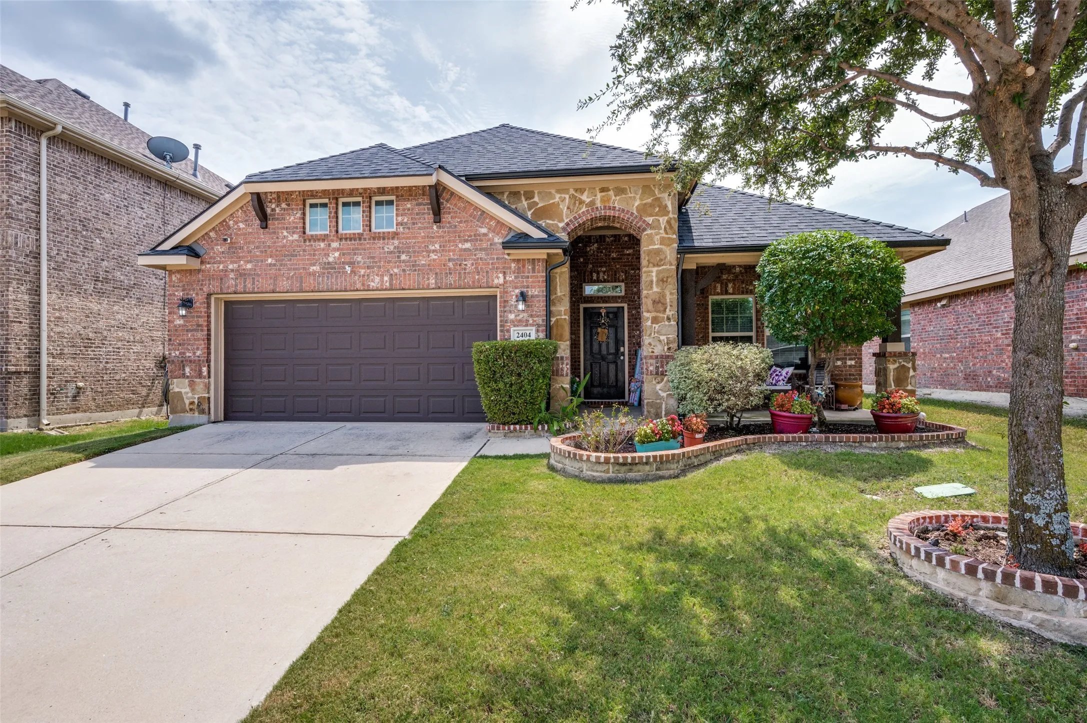 Brick and stone facade residence featuring an arched entry, dark panel garage door, concrete driveway, and a manicured lawn with mature trees