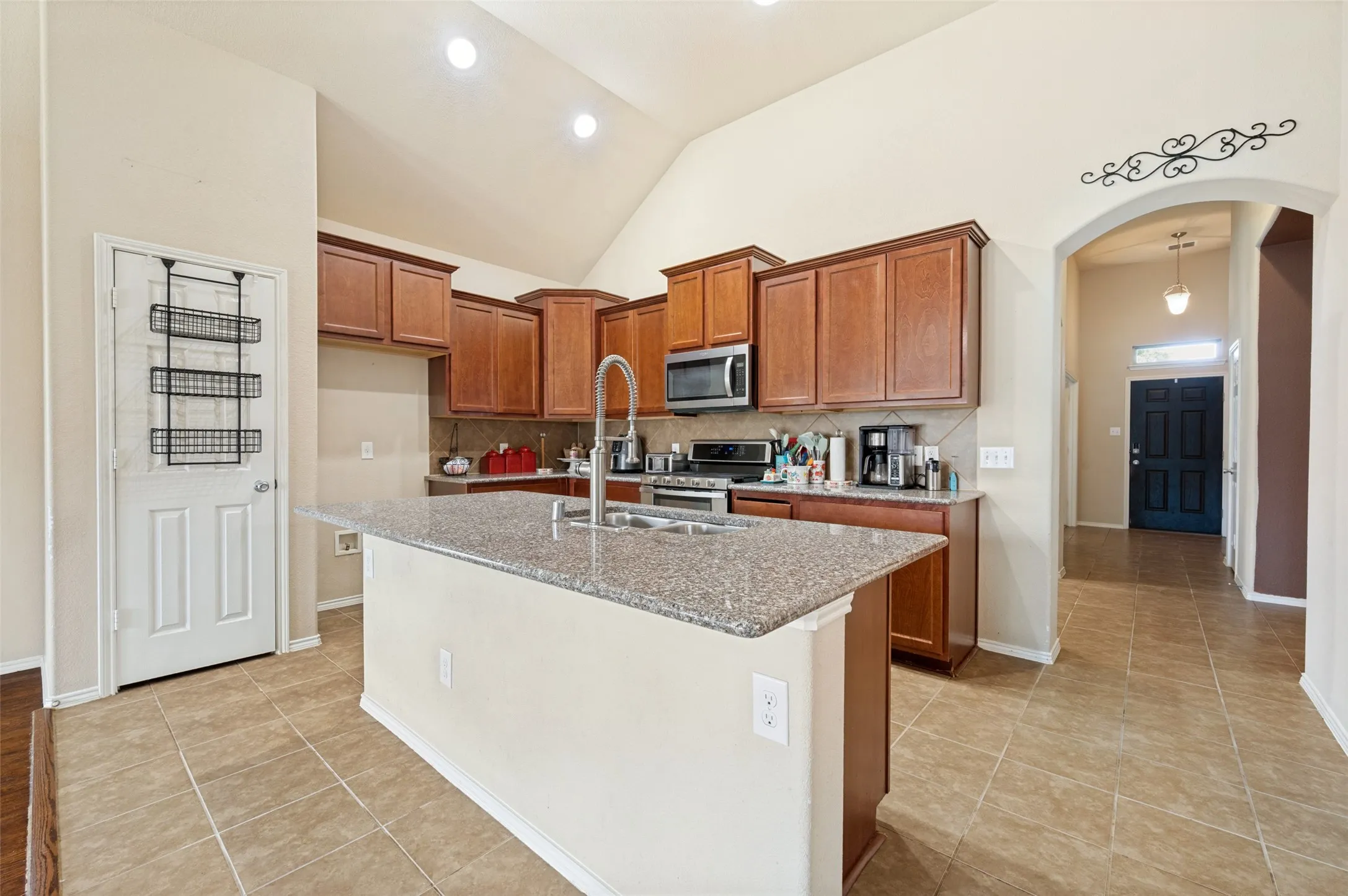 Kitchen featuring an island with a built-in sink, stainless steel appliances, wood cabinetry, tile flooring, and vaulted ceilings with recessed lighting