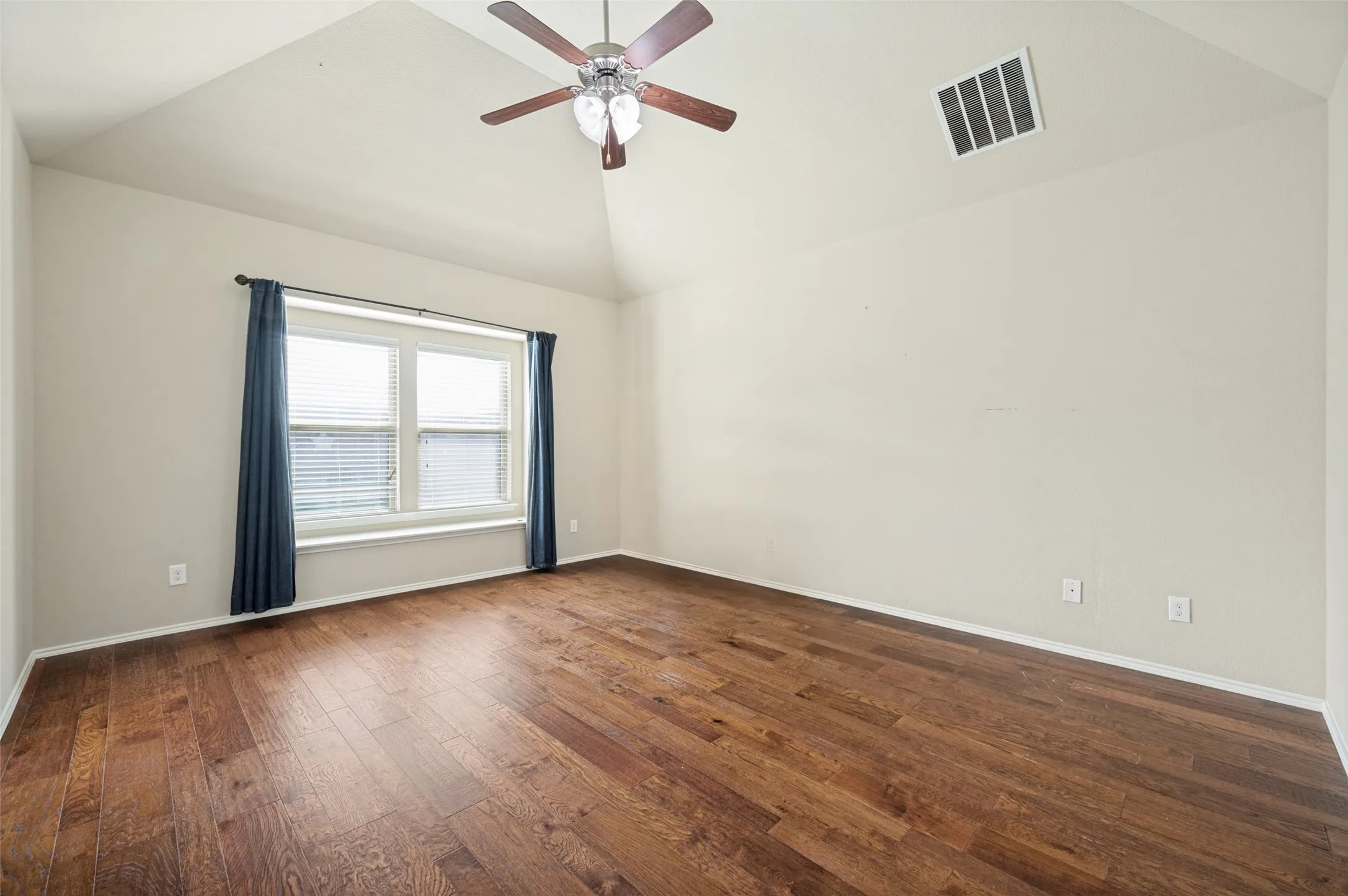 Spacious room featuring vaulted ceilings, wood-finish flooring, a ceiling fan with light fixture, and a large window with blinds