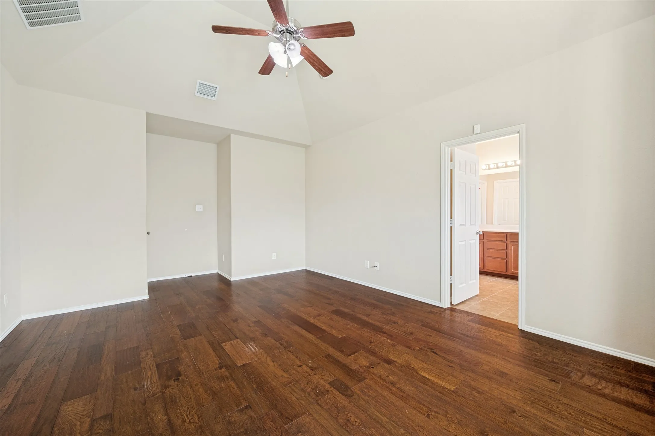 Spacious room featuring vaulted ceilings, wood-finish flooring, a ceiling fan, and direct access to a bathroom with a vanity and tile flooring