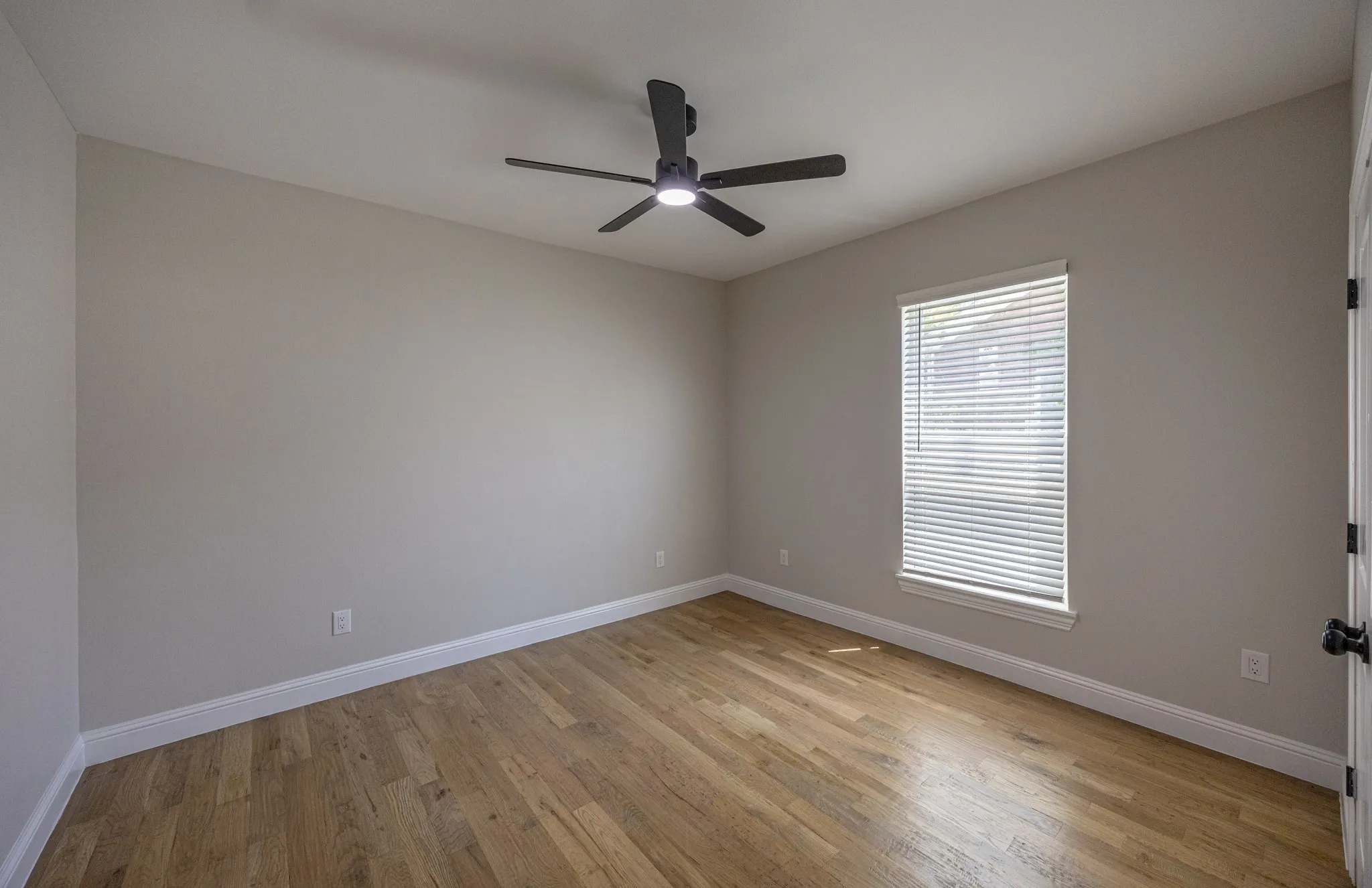Spacious room featuring wood-finish flooring, light neutral wall tones, white baseboards, a single window with blinds, and a modern black ceiling fan with integrated lighting