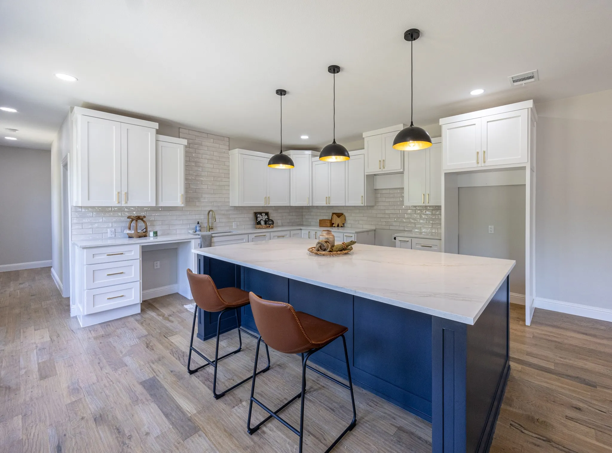 Modern kitchen featuring a large central island with a contrasting navy base and light-toned countertop, white cabinetry with gold-toned hardware, light brick-patterned backsplash, and wood-finish flooring