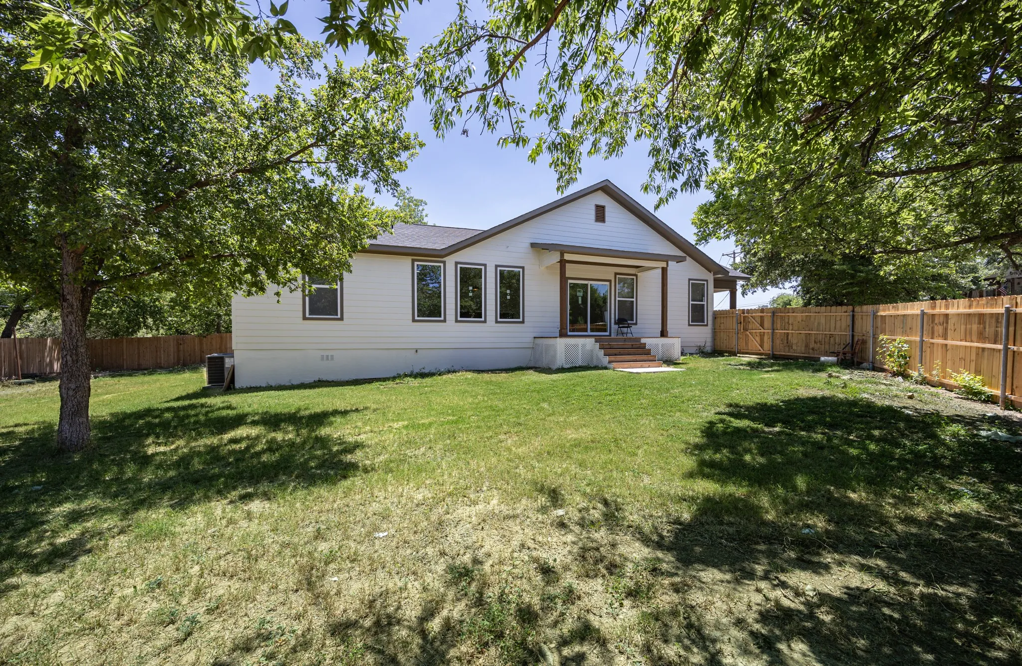 Expansive backyard featuring a green lawn, mature trees, a wooden privacy fence, and a covered patio with sliding glass doors