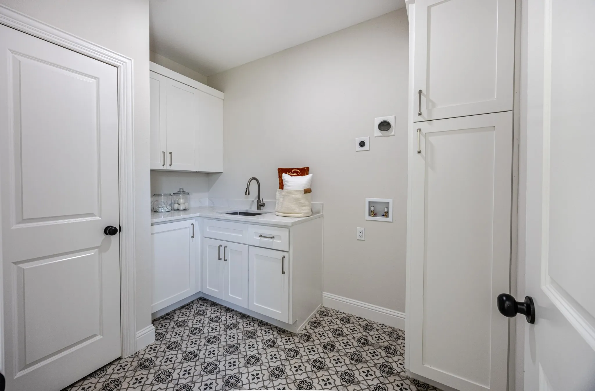Utility room featuring patterned floor tiles, white shaker cabinetry, a corner sink with a gooseneck faucet, light-toned countertops, and built-in vertical storage