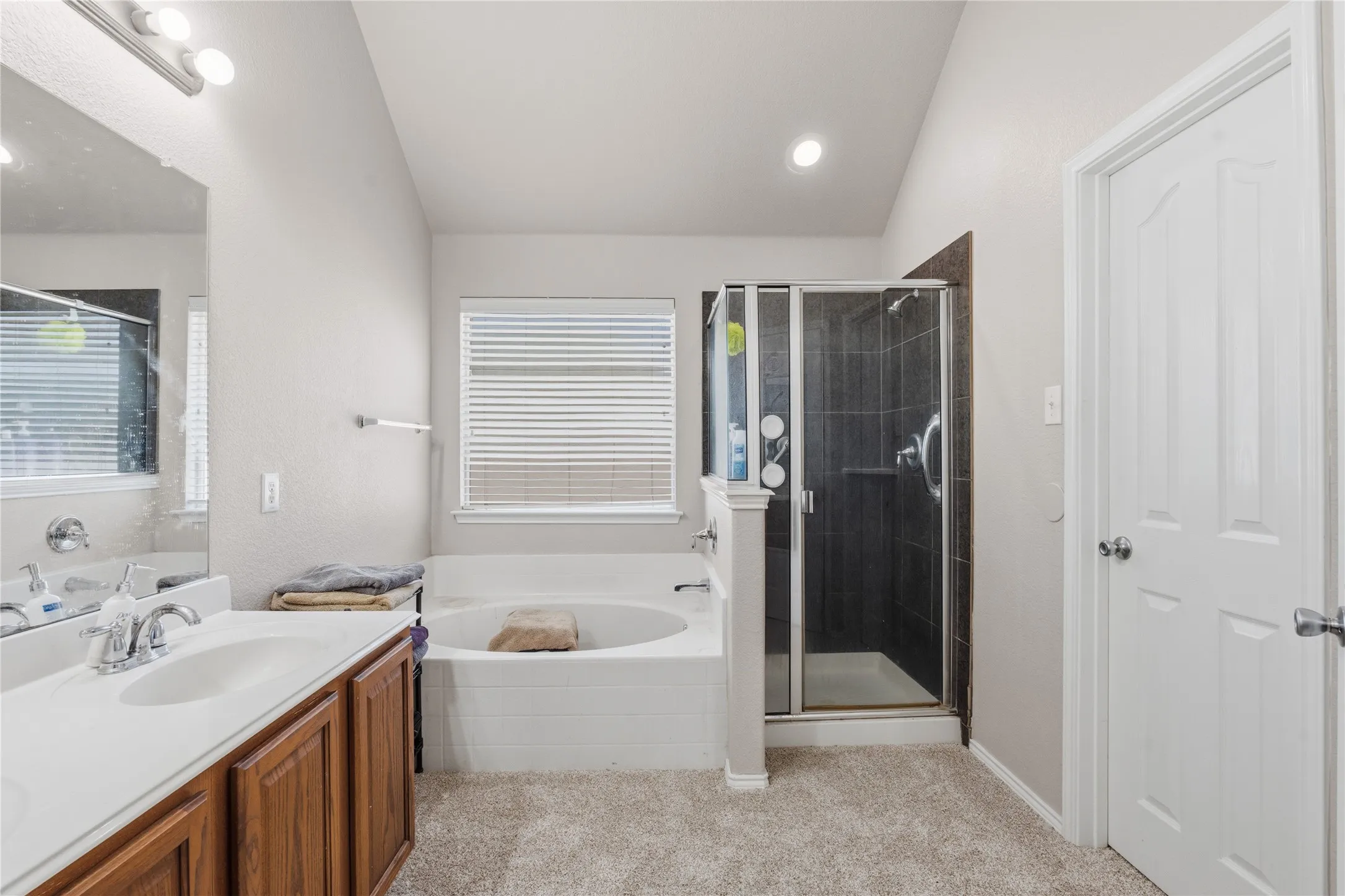 Bathroom featuring a dual vanity with a white countertop and wood-finish cabinetry, a built-in soaking tub, a separate shower with dark tile surround and glass enclosure, and recessed lighting