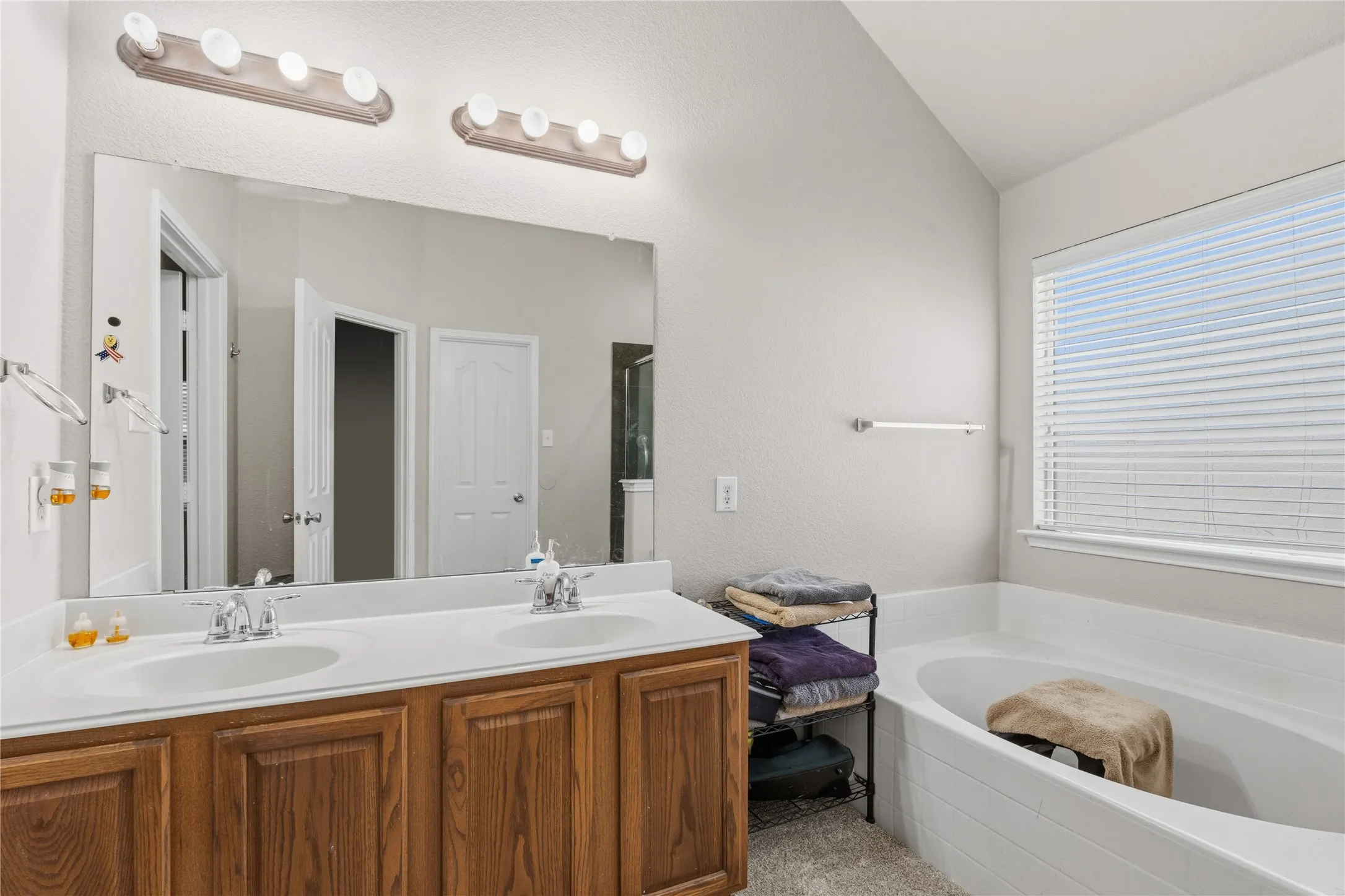 Bathroom featuring a dual vanity with wood-finish cabinetry, a large integrated mirror, overhead bar lighting, and a built-in soaking tub with tiled surround
