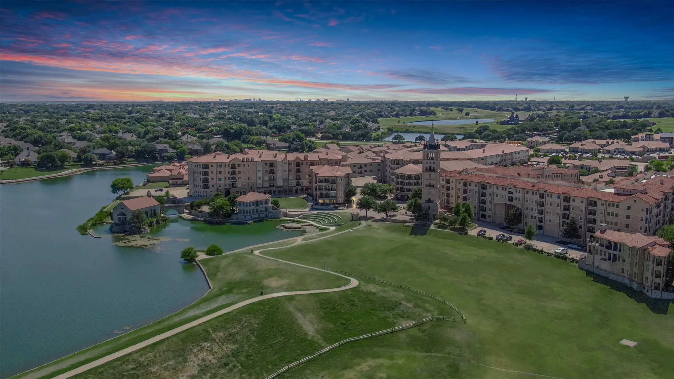 Extensive green space bordering a body of water, surrounded by multi-story structures featuring stone facades and terracotta-style roofing