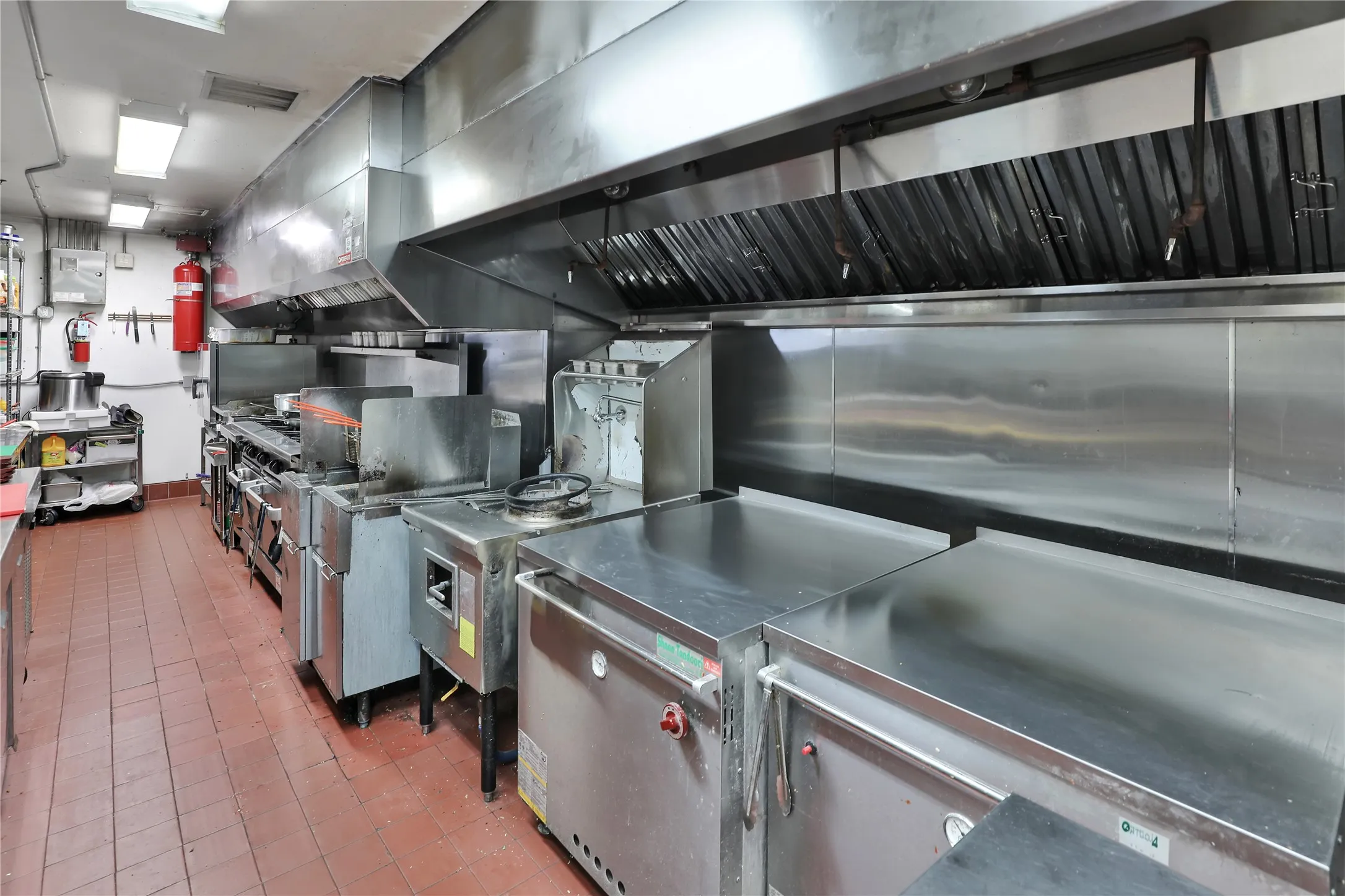 Commercial kitchen featuring stainless steel equipment, expansive ventilation hoods, and red tile flooring