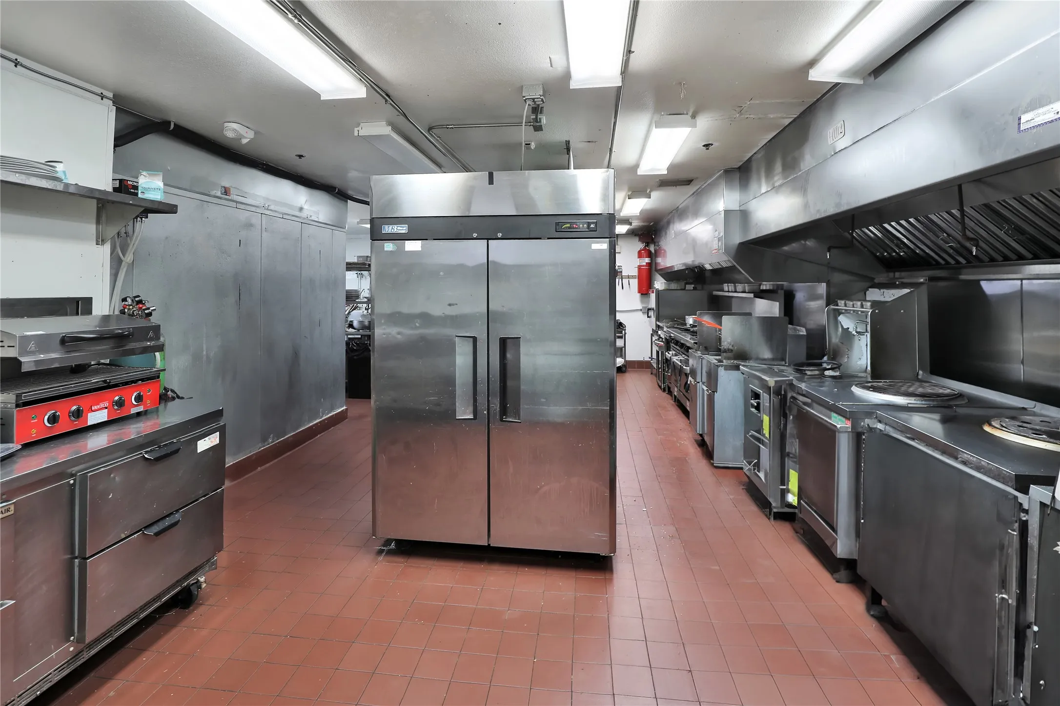 Commercial kitchen featuring extensive stainless steel appliances, red tile flooring, and overhead fluorescent lighting