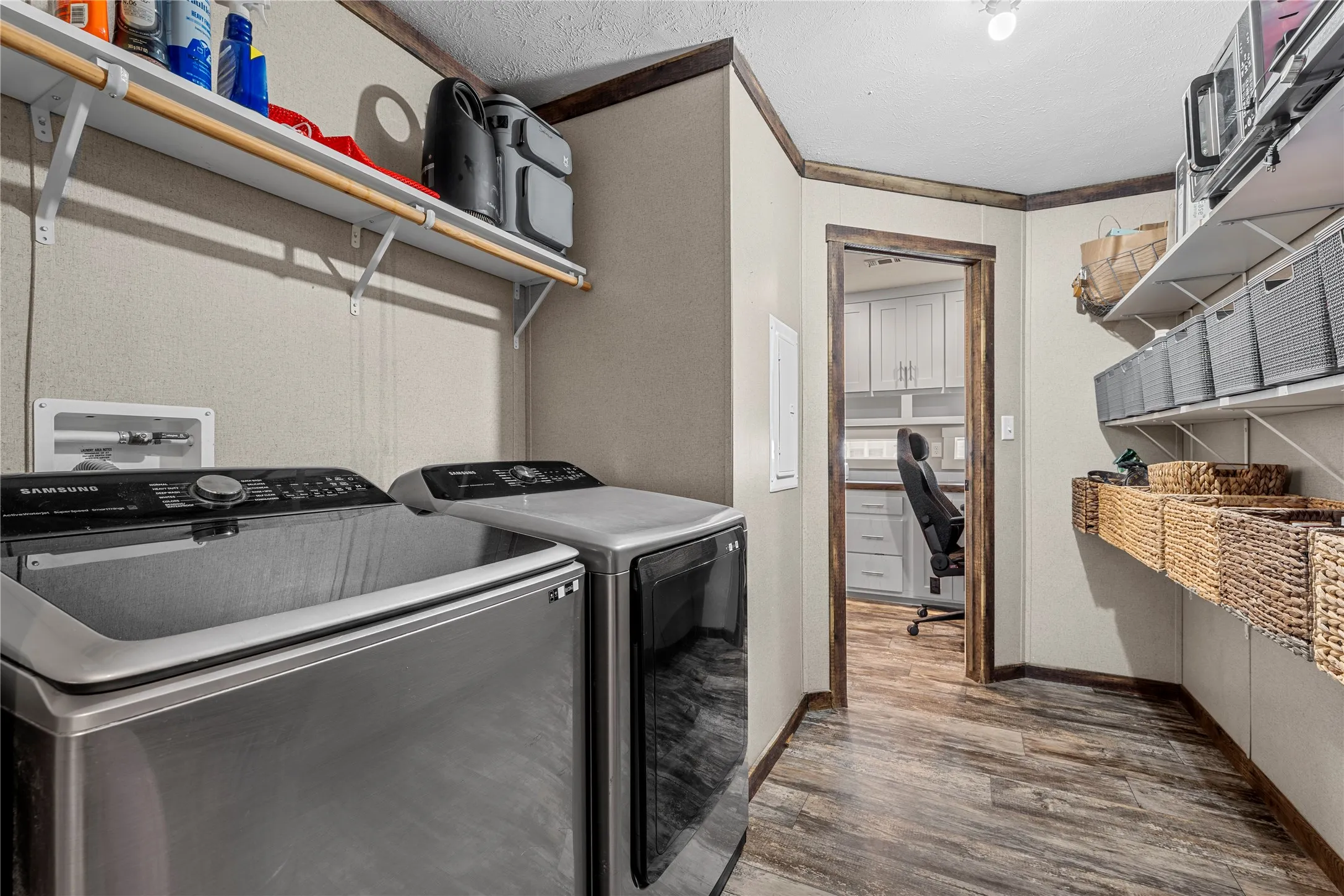 Dedicated laundry area featuring wood-finish flooring, built-in shelving, and a doorway opening to an additional room with white cabinetry