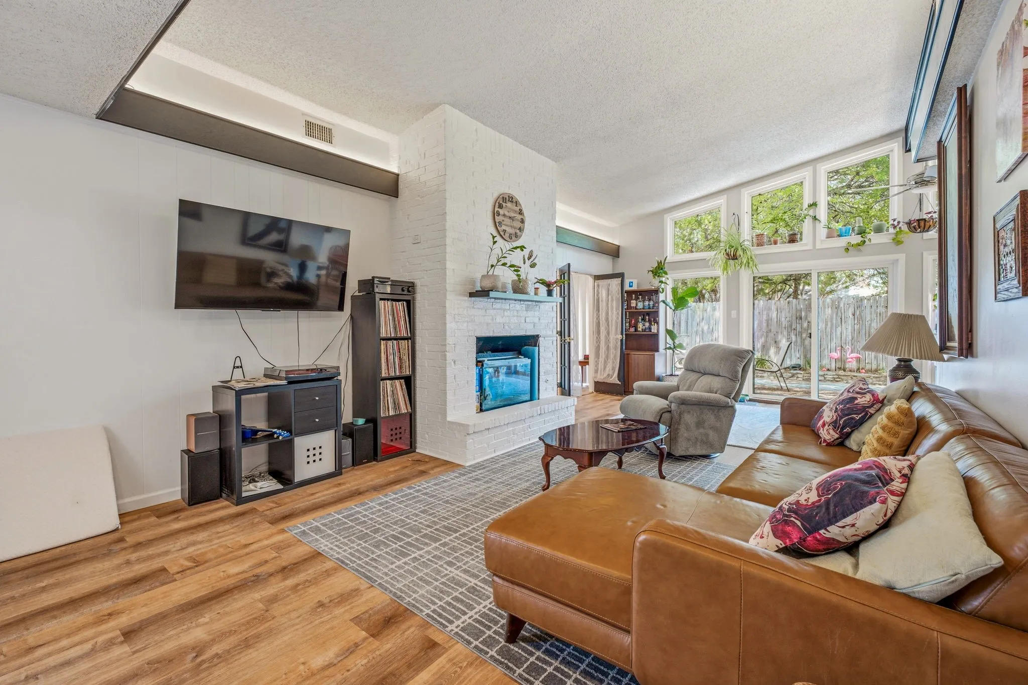 Spacious living area featuring wood-finish flooring, a white brick fireplace, and extensive natural light from multiple large windows and sliding glass doors