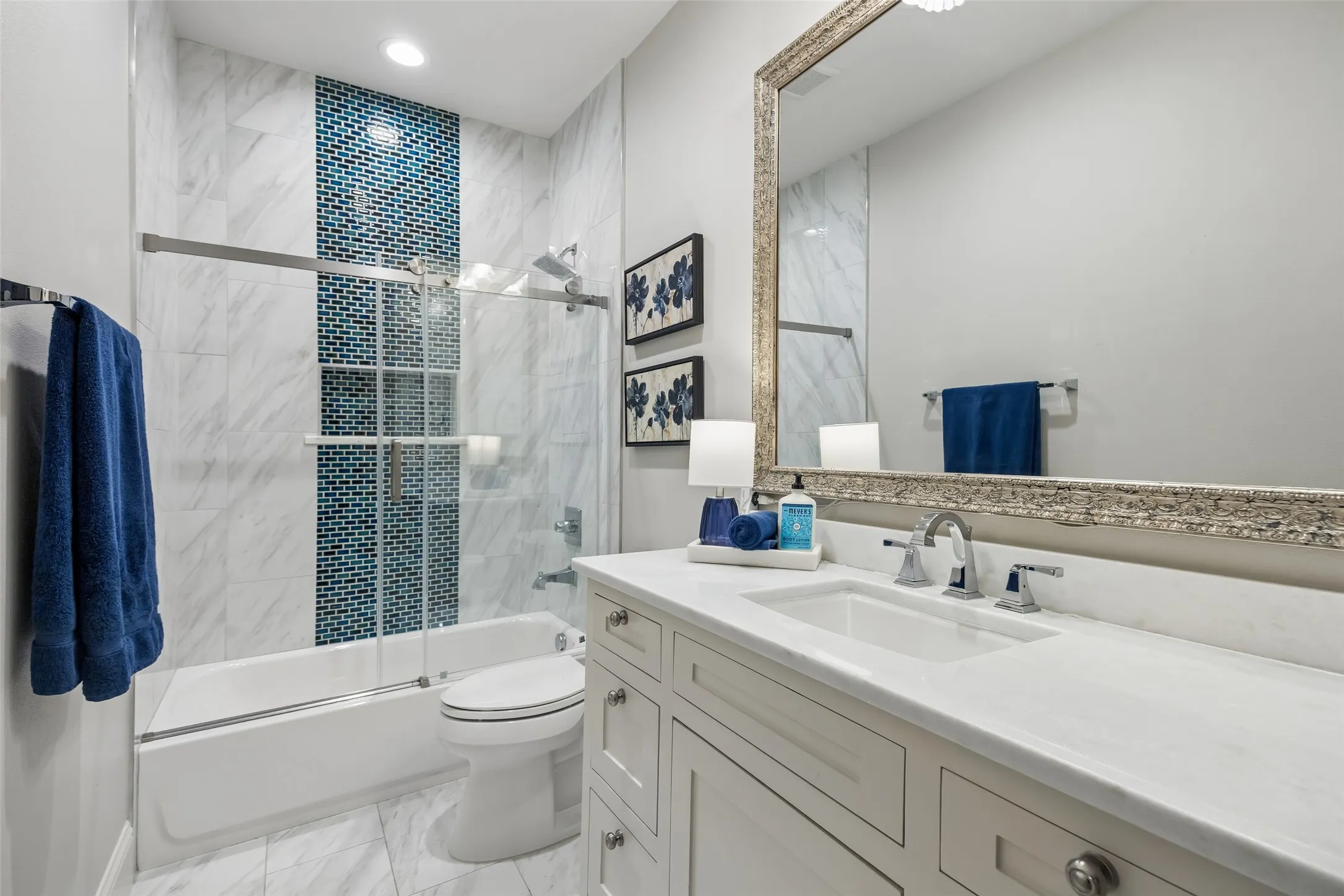 A Bright Full Bath with Bold Accent Tile – The upstairs bath pairs a long vanity and quartz counter with a tub-shower surround featuring marble-look tile and a vertical blue mosaic accent.