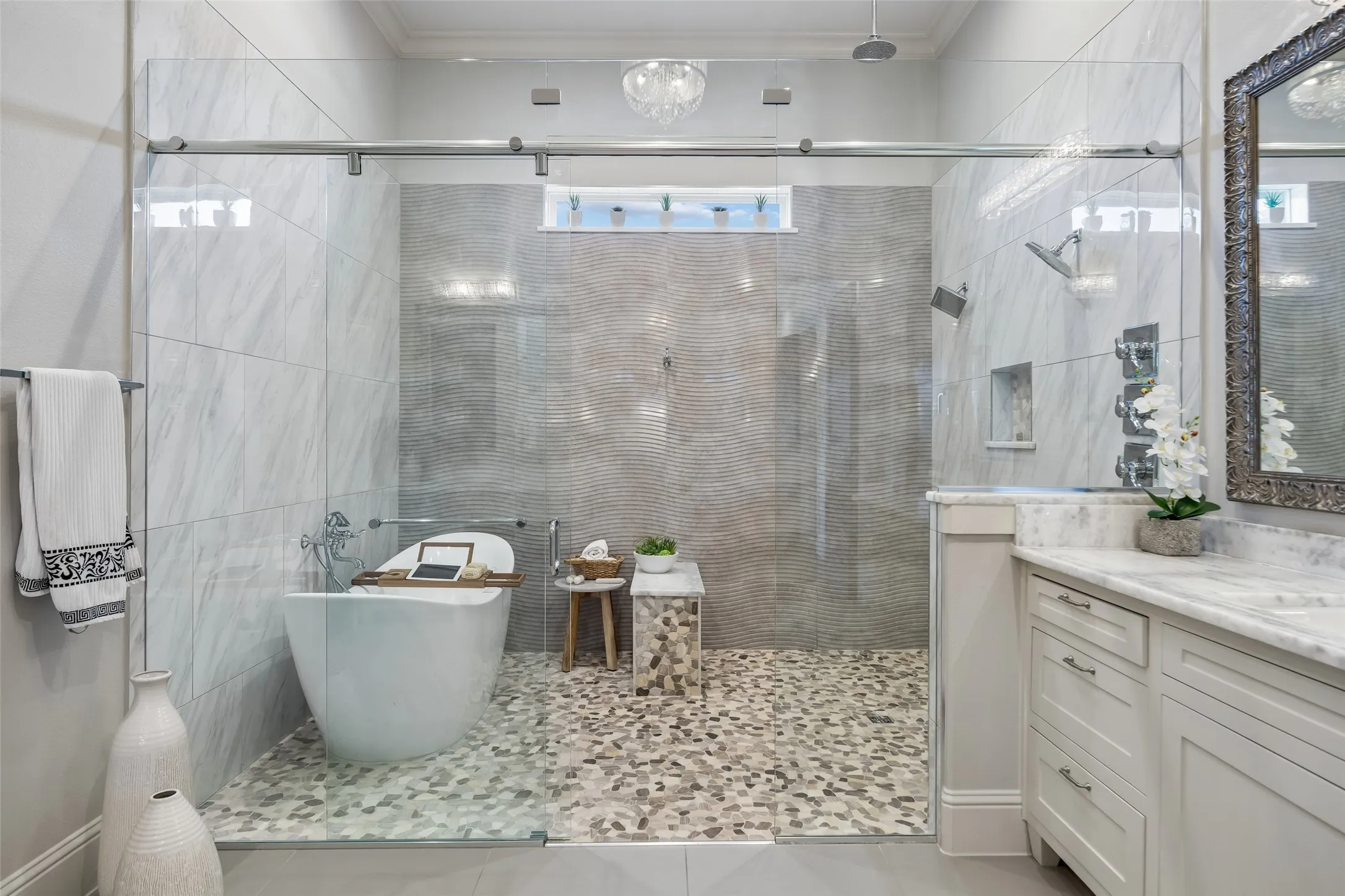 A Wet Room Designed for a True Spa Experience – The oversized glass enclosure brings together the freestanding soaking tub, dual shower stations, handheld fixtures, pebble flooring, and textured tile in one dramatic bathing space.