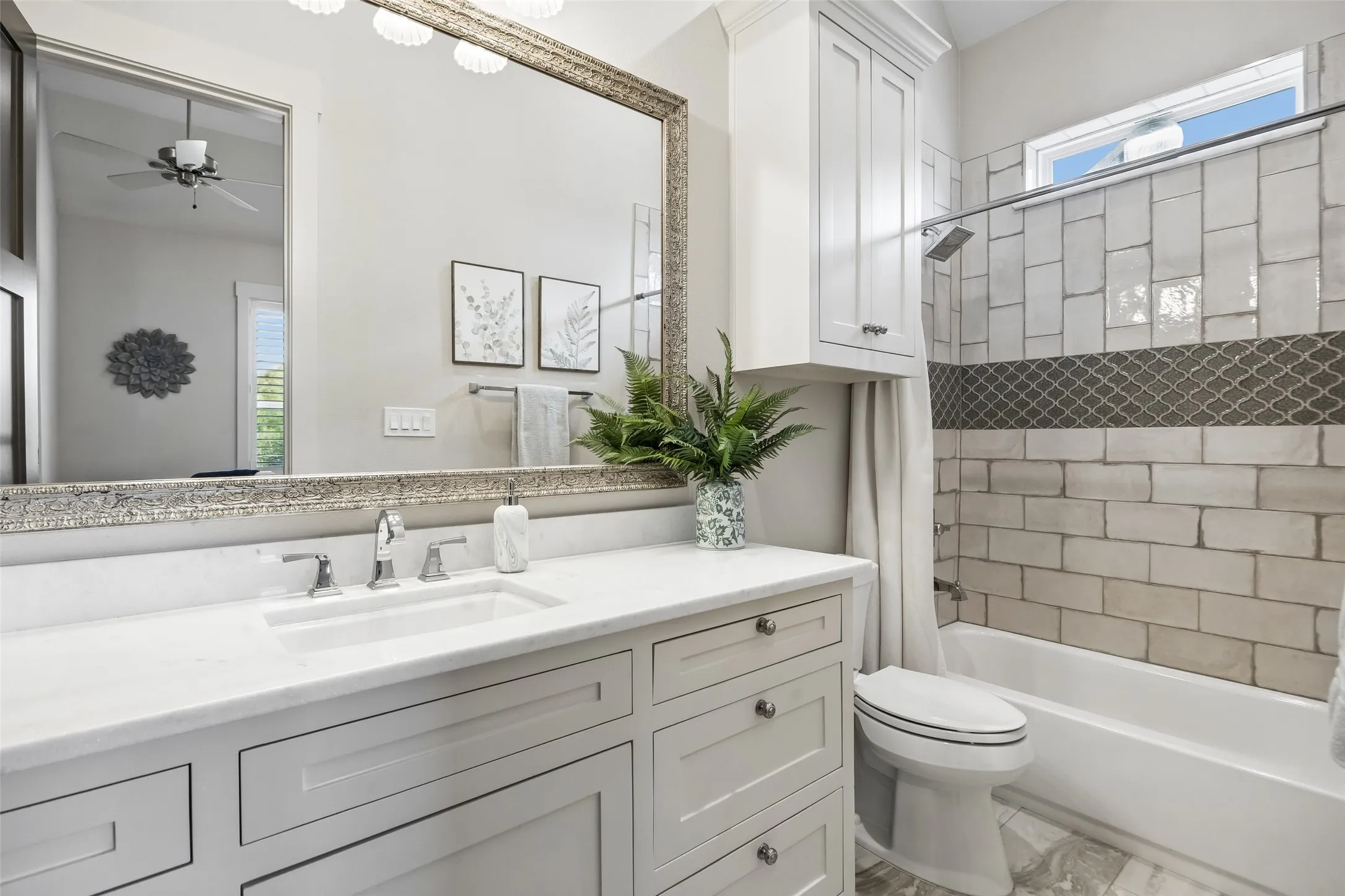 A Private Bath with Custom Tilework – The ensuite bath features a long quartz-topped vanity, abundant cabinetry, a tiled tub-shower surround, decorative accent detail, and a high window that brings in natural light while maintaining privacy.