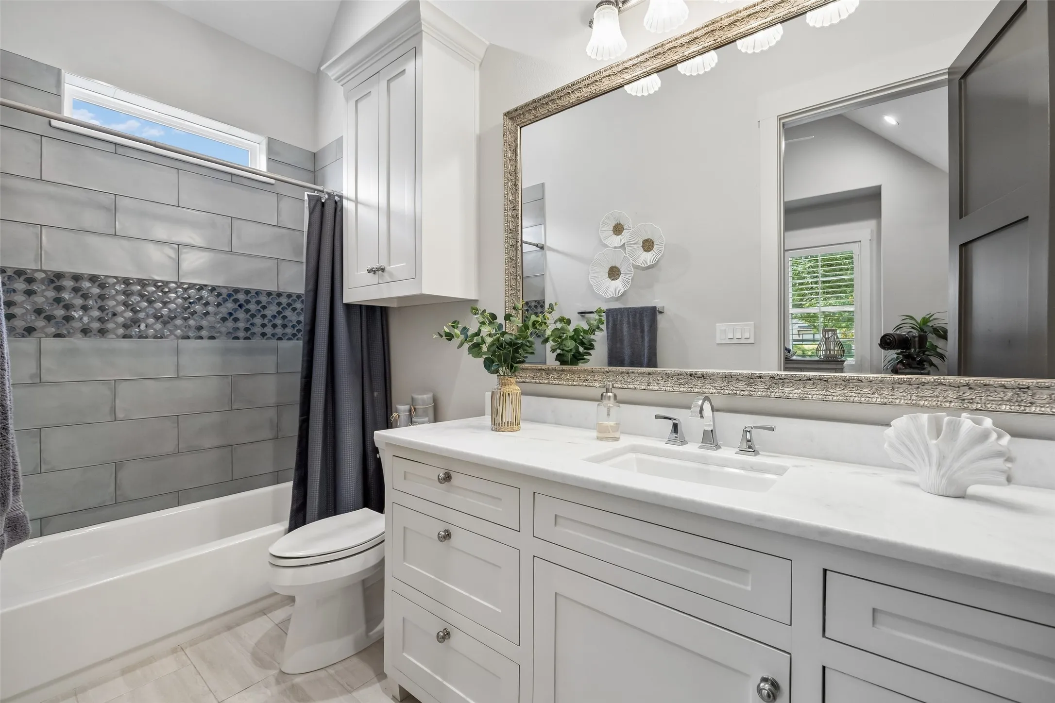 A Secondary Bath with Thoughtful Finishes – A long quartz-topped vanity provides generous counter and storage space, while the tub-shower combination features large-format tile, a decorative accent band, and natural light from the high window.