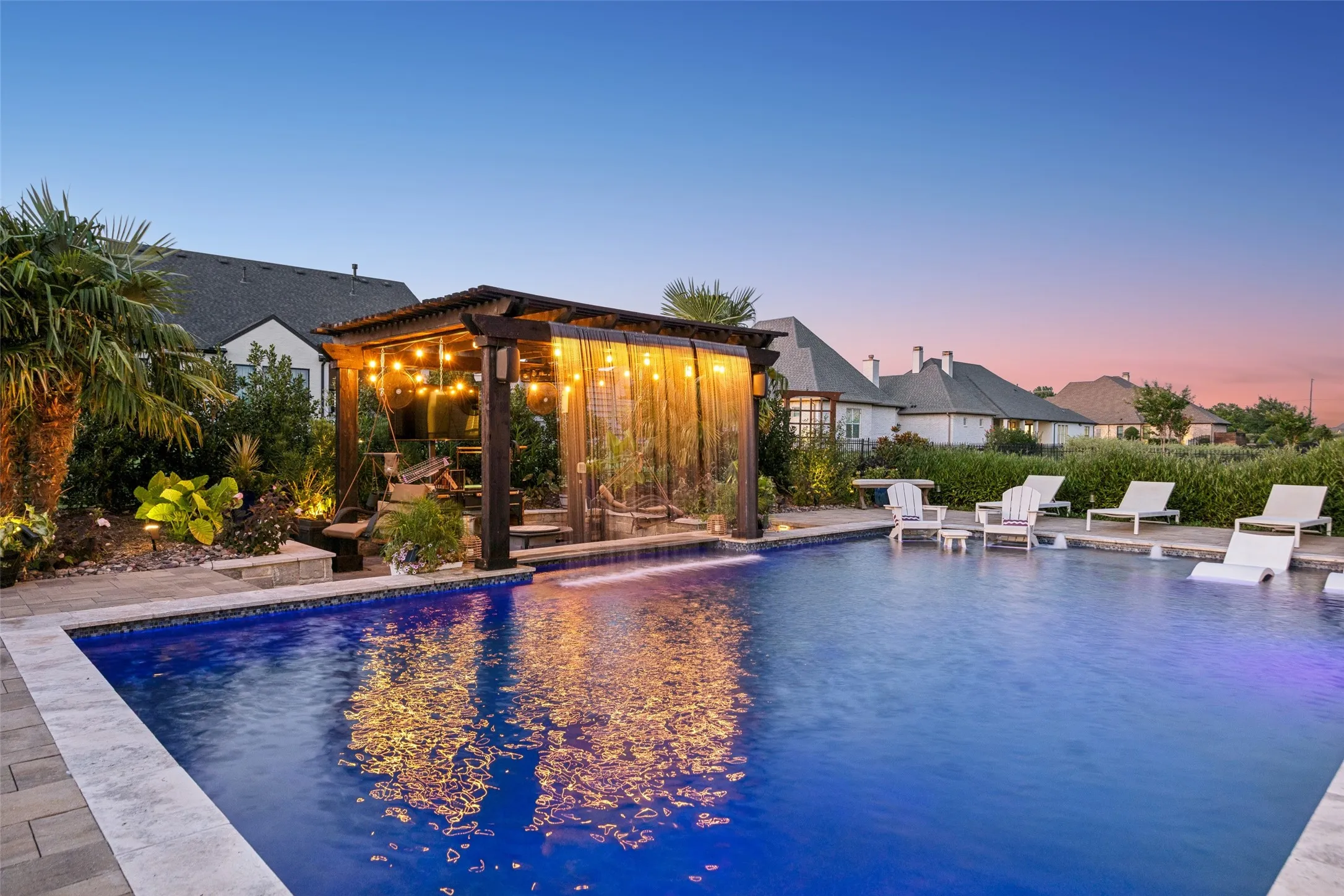 A Backyard That Steals the Show – The spacious resort-style pool offers a broad swimming area, in-water loungers, shallow seating, swim-up bar seating, and cascading water features, all framed by pavers, lush landscaping, and a warmly lit pergola that turns this private retreat into an unforgettable destination from sunset well into the night.