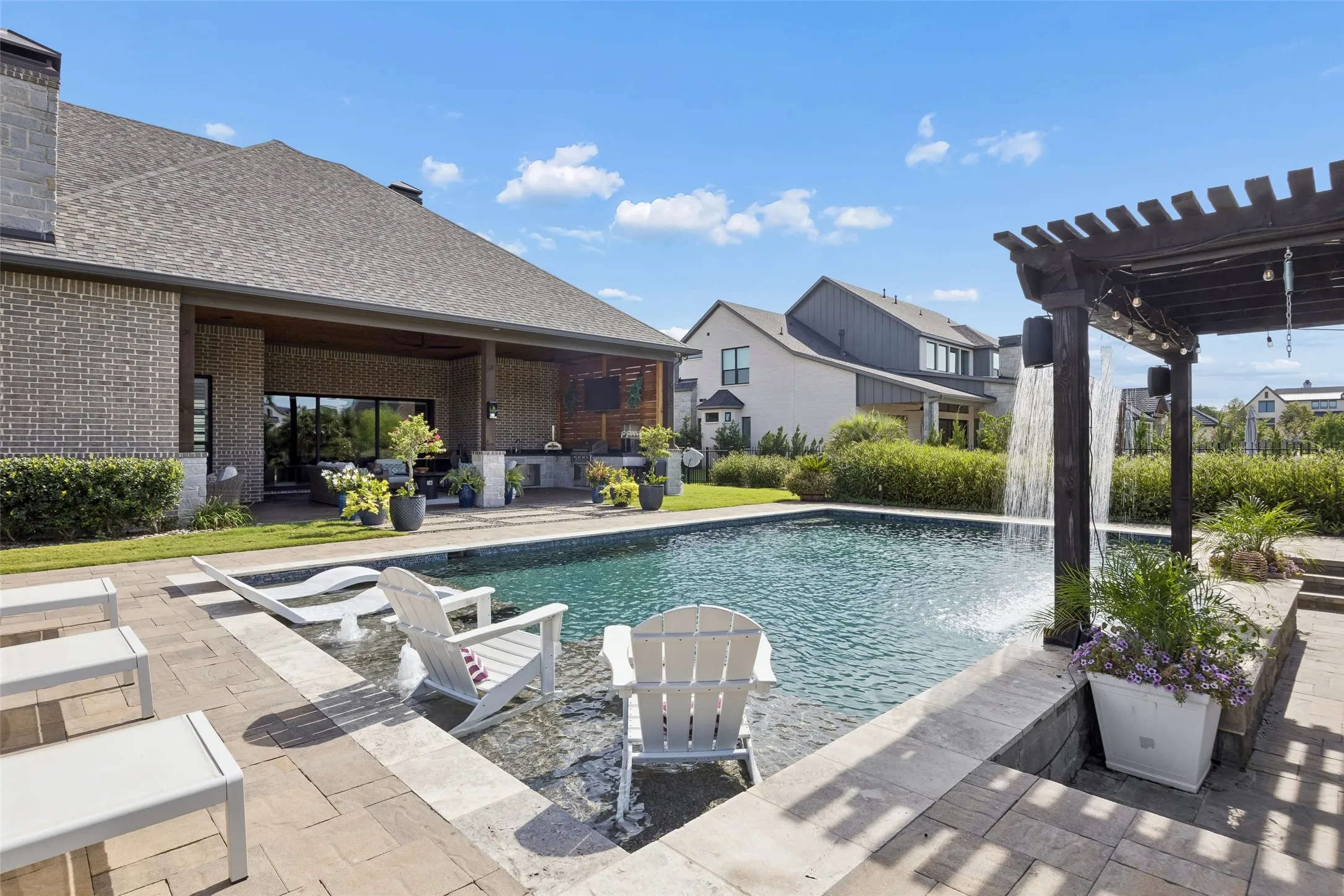A Pergola Retreat Beside the Water – The cedar pergola adds another shaded destination with hanging seating, string lights, fans, and a dramatic curtain of water along the pool’s edge.