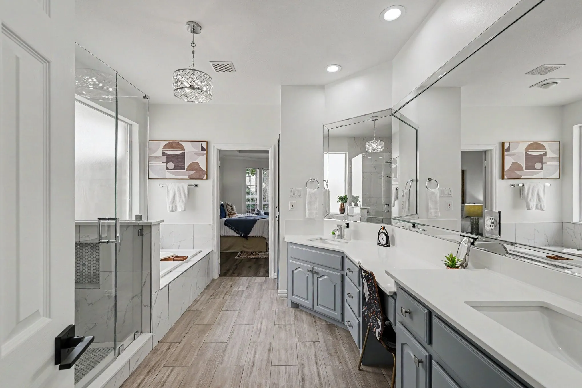 Spacious bathroom featuring a frameless glass shower enclosure, a built-in soaking tub, wood-finish flooring, dual vanities with light gray cabinetry, and an expansive wall-mounted mirror