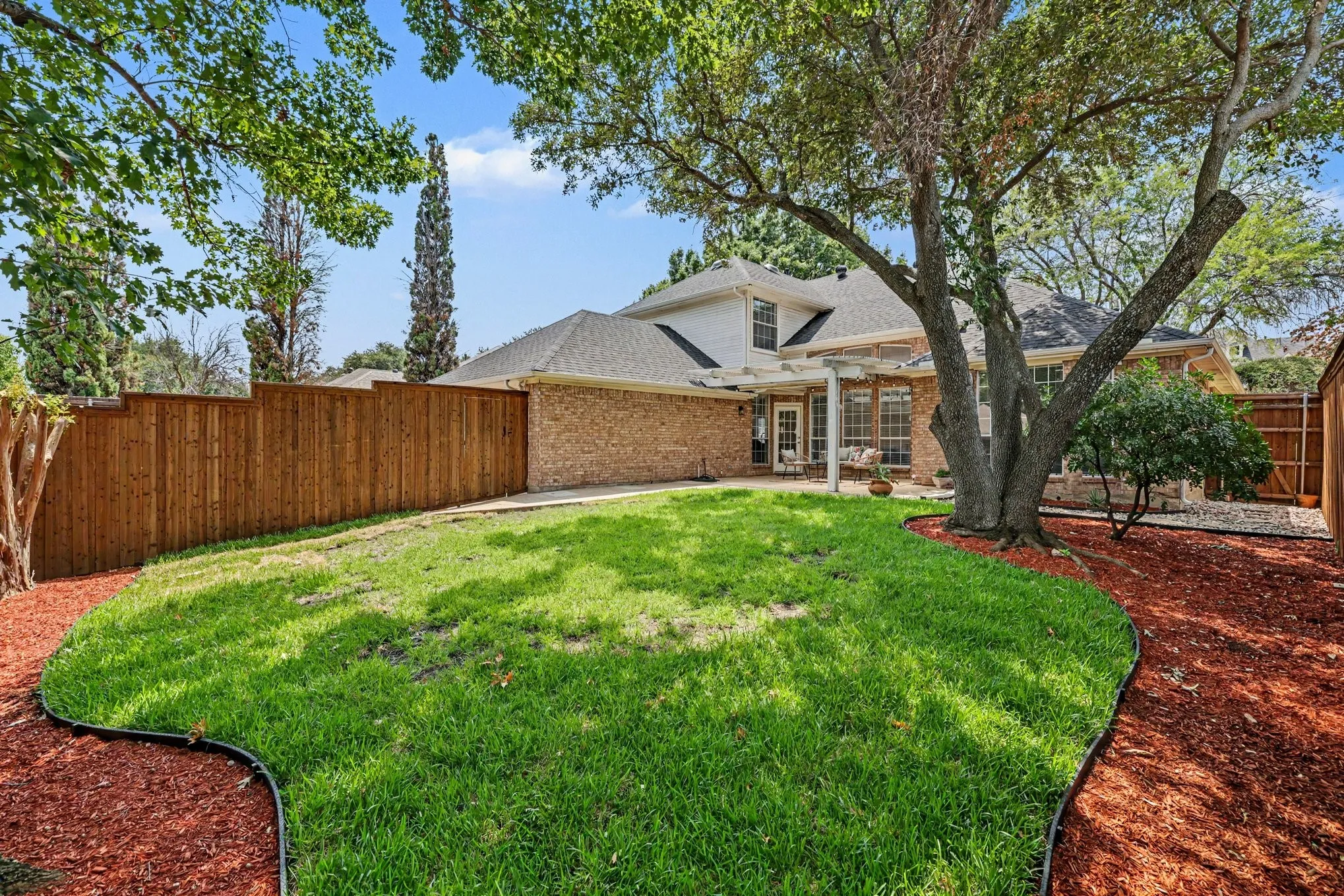 Expansive fenced backyard featuring a manicured lawn, established shade trees, and a mulched perimeter
