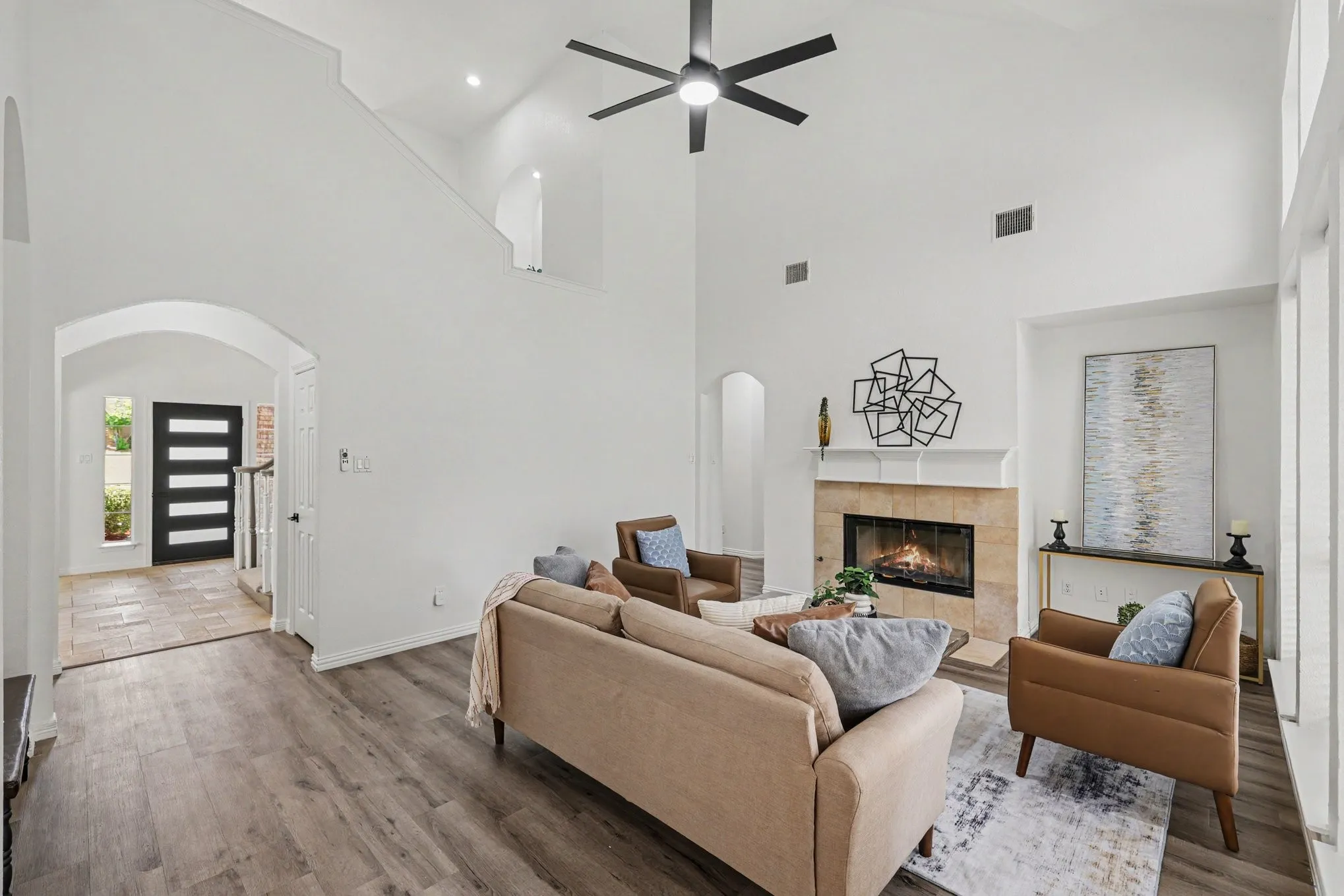 Spacious living room featuring vaulted ceilings, a modern ceiling fan, wood-finish flooring, a tiled fireplace with mantel, and an arched entry to the foyer