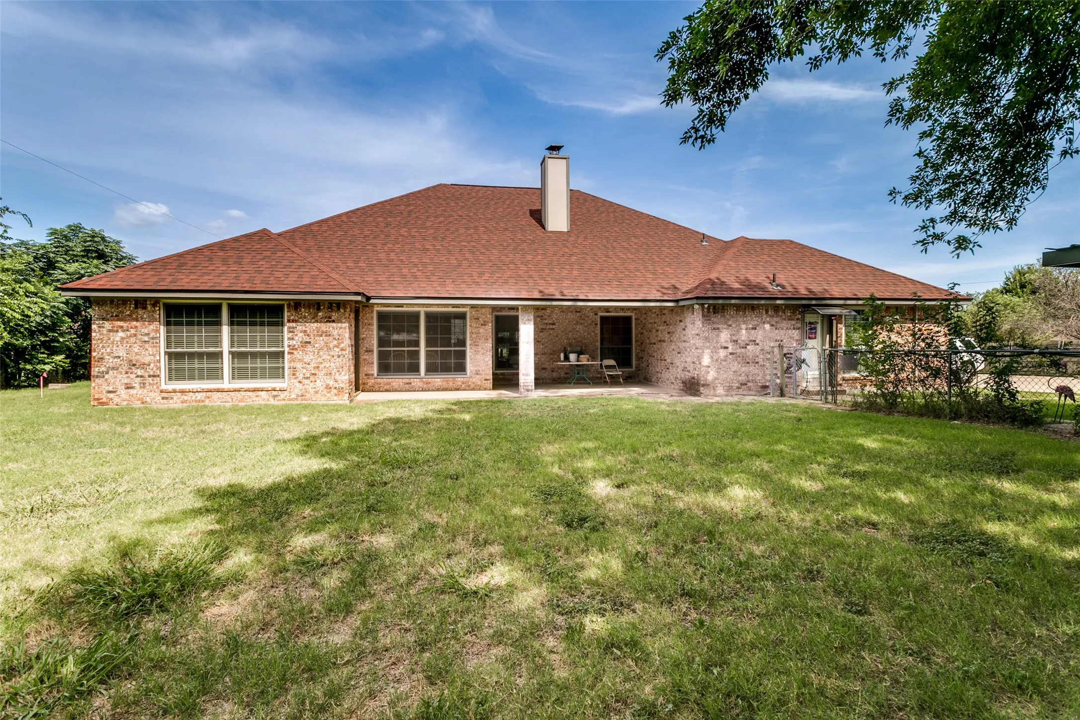 Expansive backyard featuring a brick exterior, a covered patio area, a chimney, and a brown shingle roof