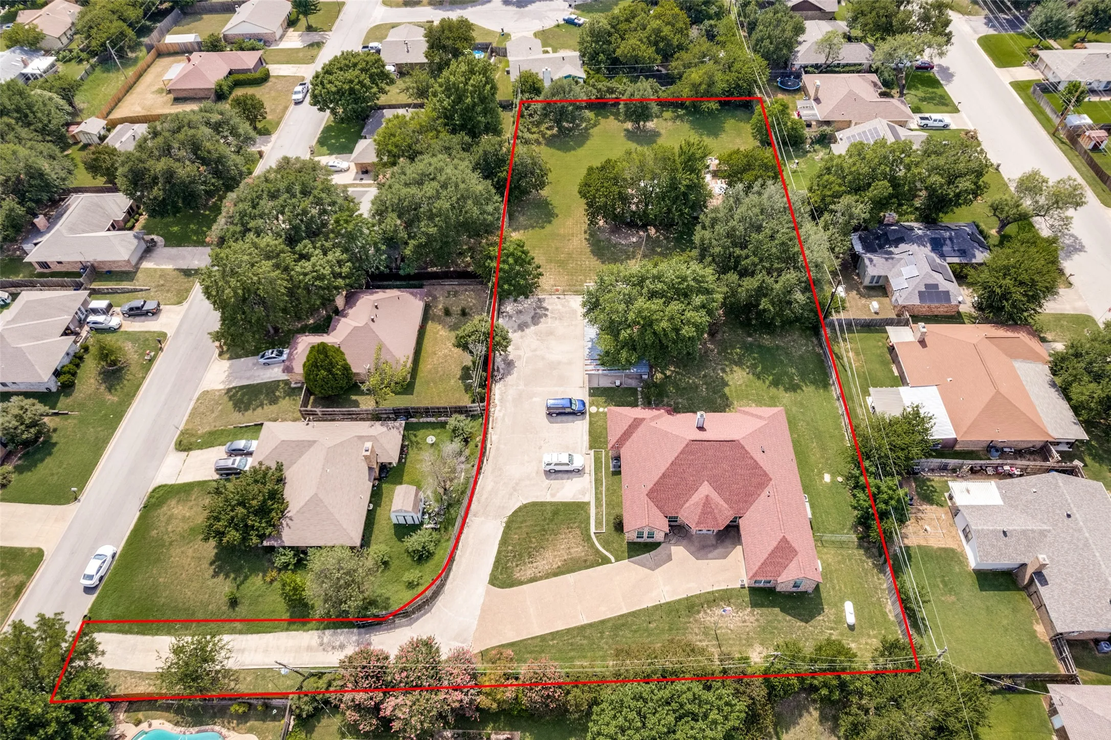 Expansive property featuring a large residence with a red shingle roof, a substantial paved driveway, and mature trees throughout the lot