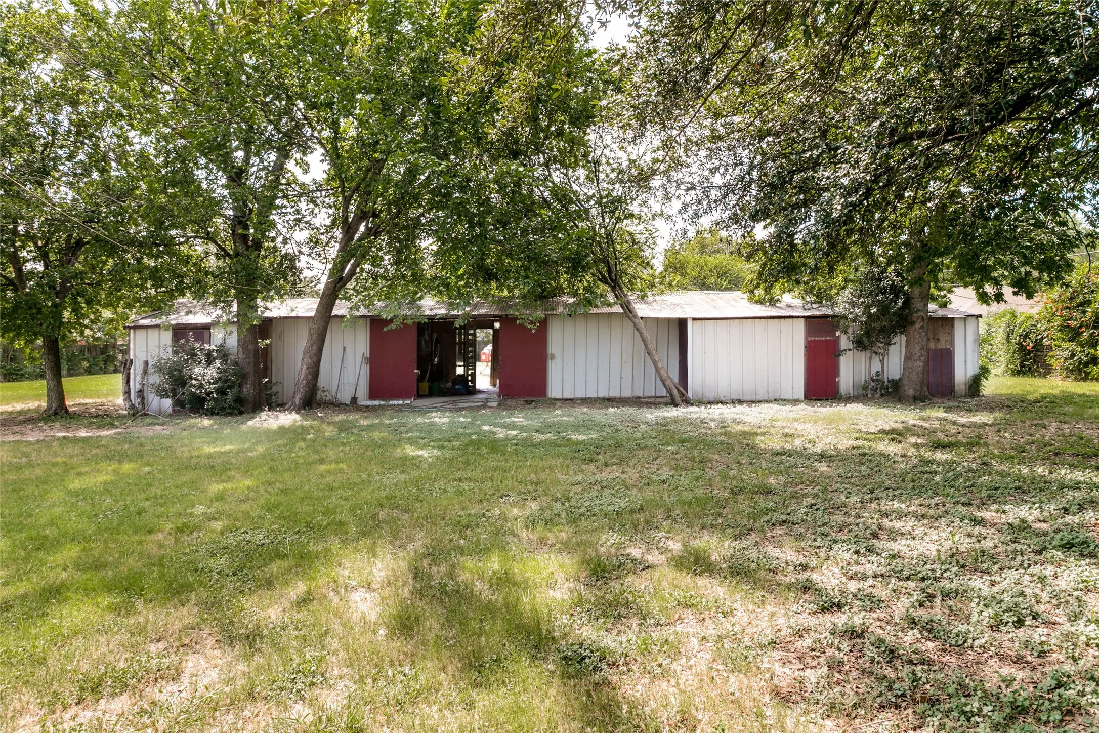 Expansive outdoor structure featuring white siding and multiple red sliding doors, situated within a spacious grassy area with mature trees