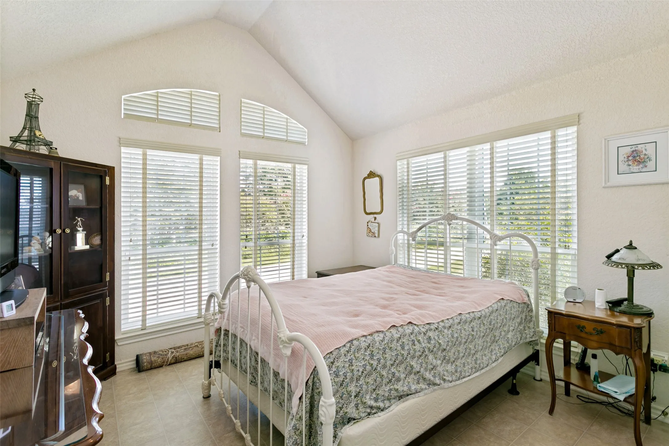 Vaulted ceiling bedroom with tiled flooring, featuring a large window array with white blinds, and arched transom windows