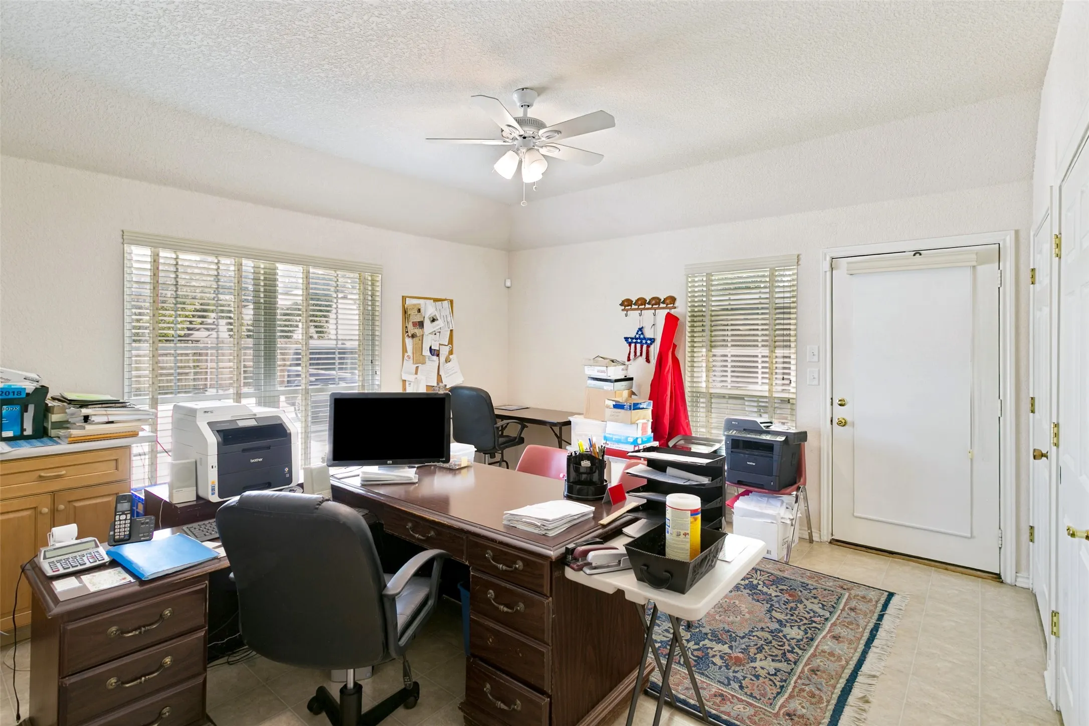 Spacious interior room featuring a vaulted ceiling with a ceiling fan, large windows with blinds, a white paneled door, and light-colored tile flooring