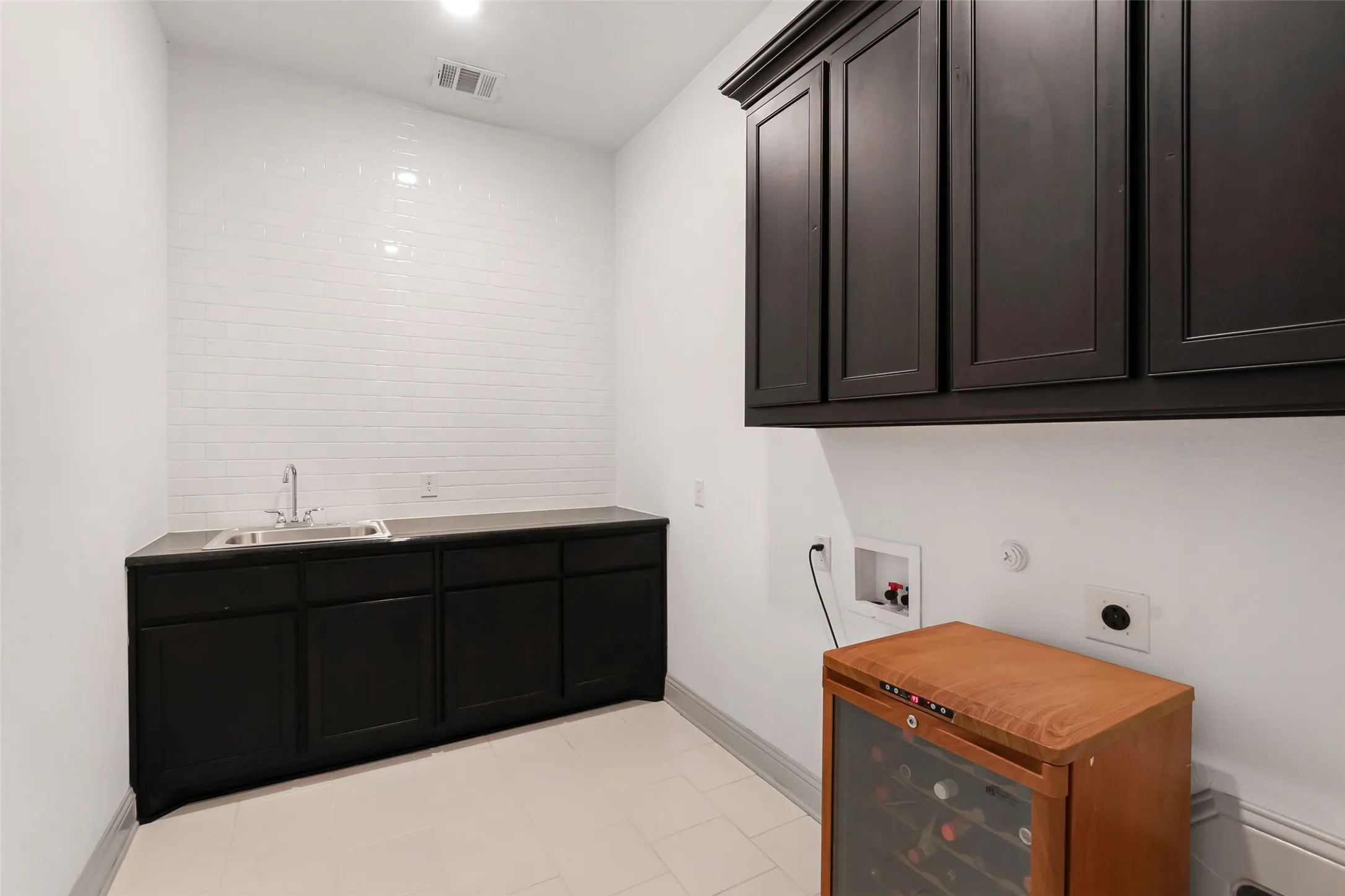 Utility area featuring white subway tile backsplash, dark wood-finish cabinetry, a stainless steel sink, and light tile flooring