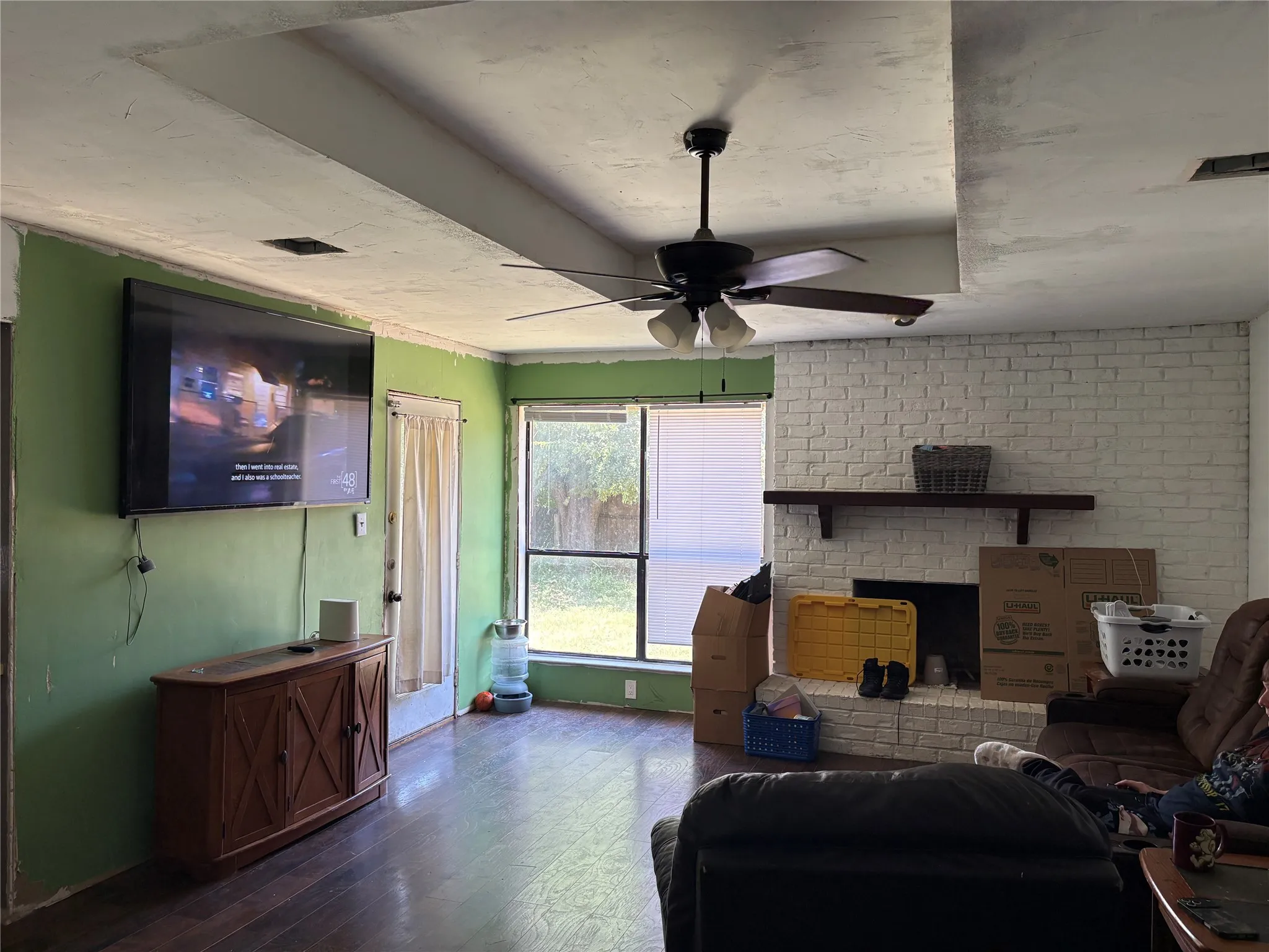 Living area featuring wood-finish flooring, a white-painted brick fireplace with a wood mantle, a ceiling fan with integrated lighting, and a large window overlooking the backyard