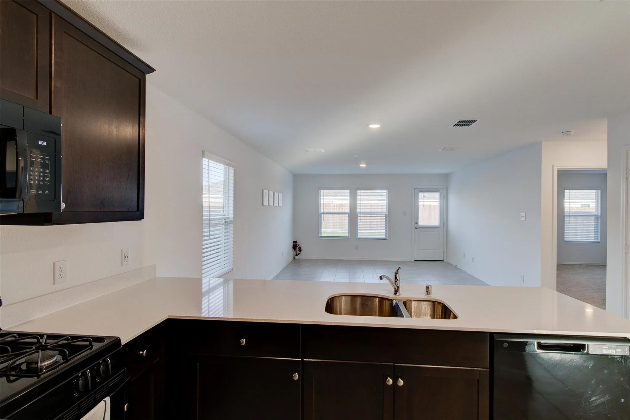 Kitchen featuring dark wood cabinetry, light-toned countertops, a built-in stainless steel double sink, and integrated black appliances including a microwave, gas range, and dishwasher